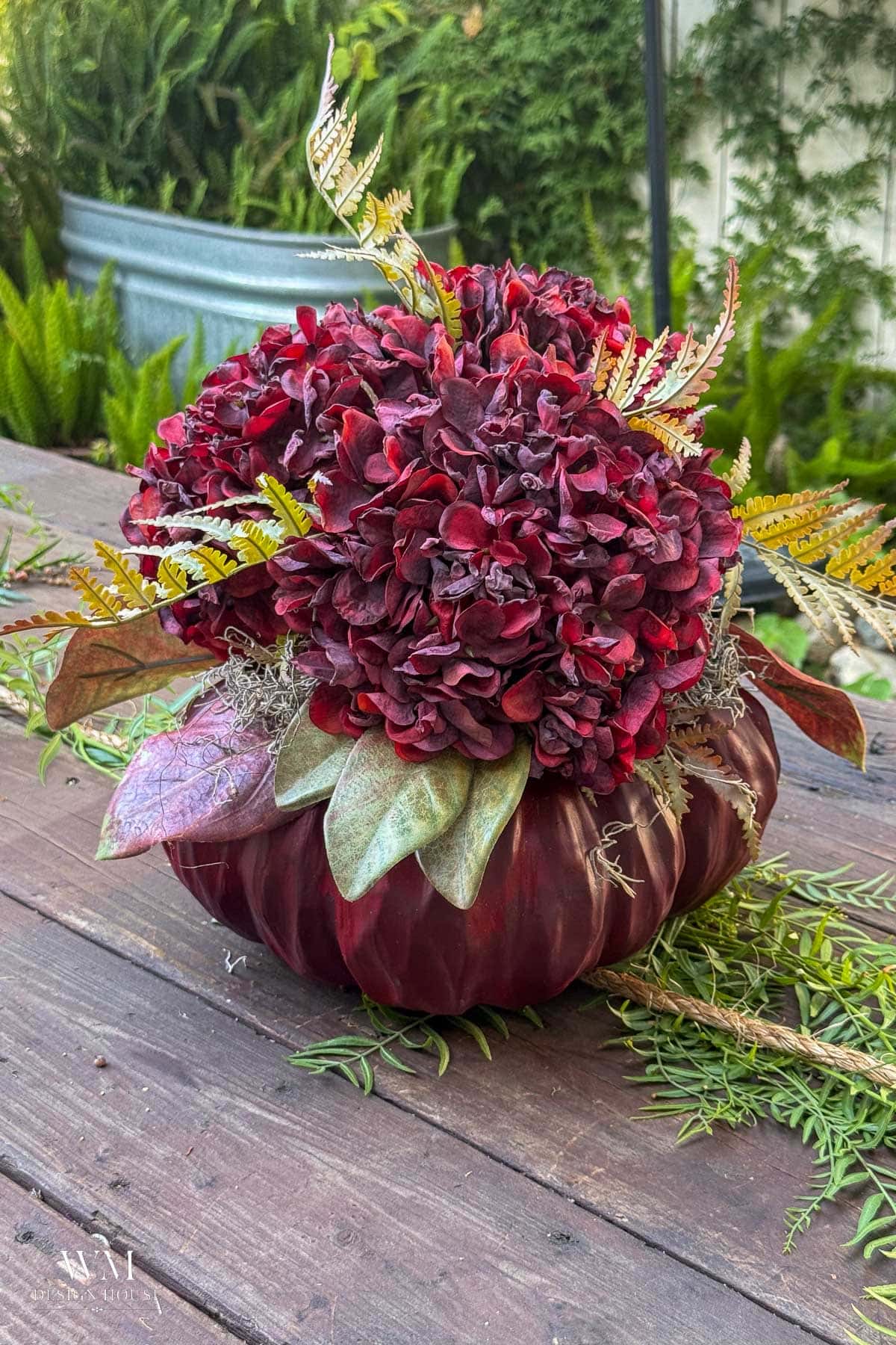 A dark red pumpkin is topped with deep red hydrangea blooms, decorative leaves, and ferns, displayed on a wooden table outdoors with greenery in the background.