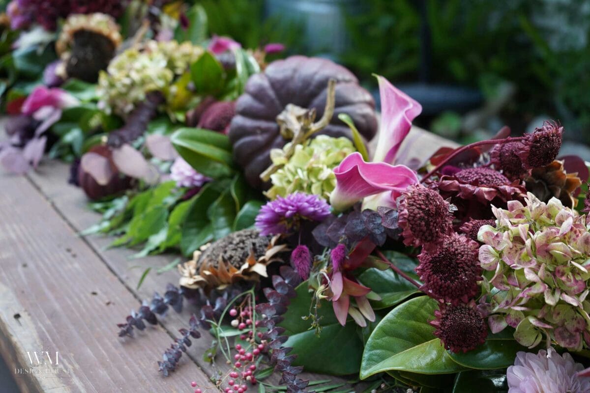 Pink calla lilies, dried burgundy dahlias, antique green hydrangeas, and a plum pumpkin tucked into a greenery table runner