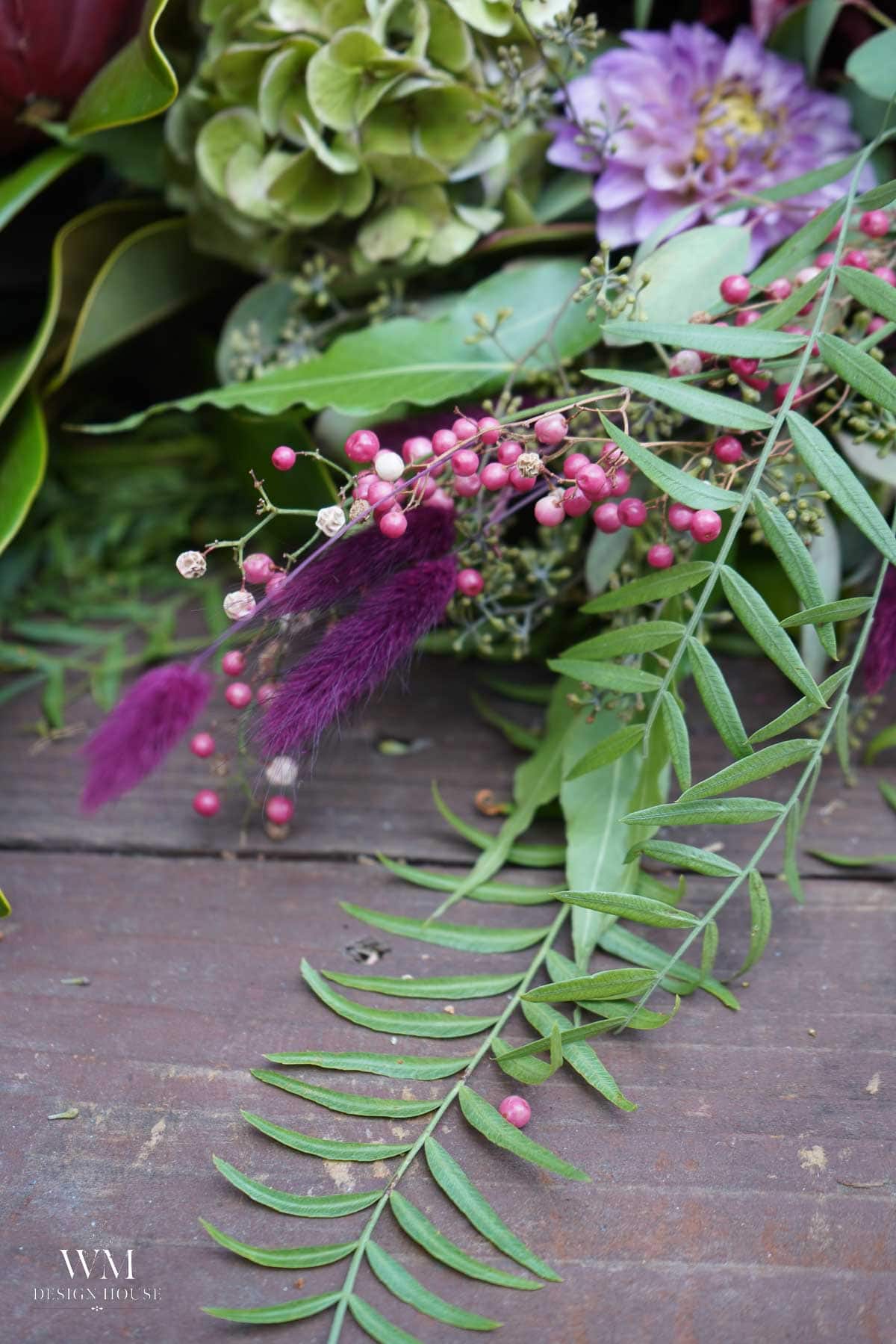 A close-up of a floral arrangement with pink berries, purple flowers, feathery accents, and green leaves, displayed on a wooden surface.