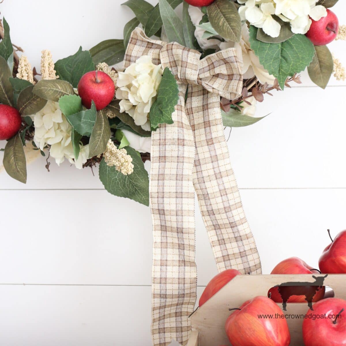 A plaid ribbon bow decorates a floral wreath with red apples and white flowers, hanging on a white wall. In the lower right, a crate holds several more red apples.