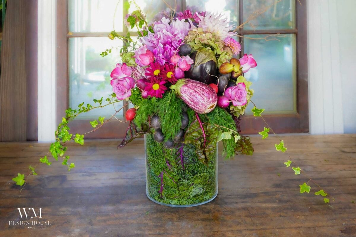 A glass vase holds a vibrant vegetable-centerpiece of pink flowers, leafy greens, and small vegetables, displayed on a wooden table in front of a window.