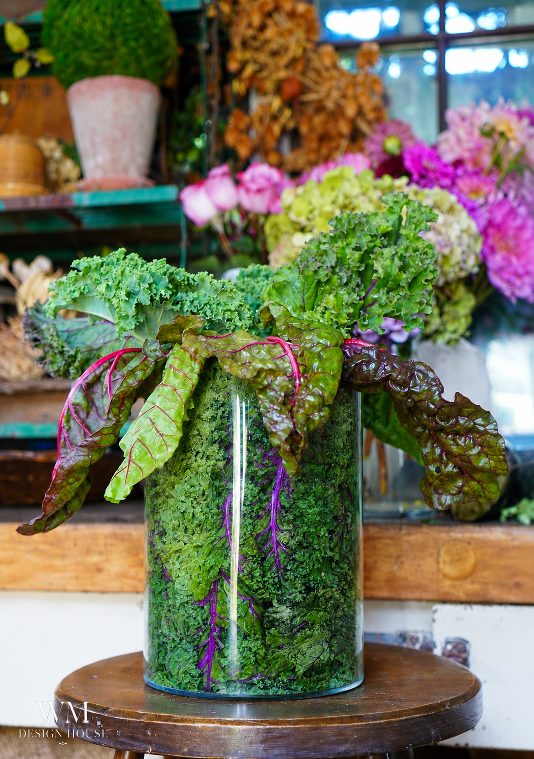 A glass vase filled with fresh green leaves, curly kale, and Swiss chard creates a stunning vegetable-centerpiece, displayed on a wooden surface with colorful flowers and plants softly blurred in the background.