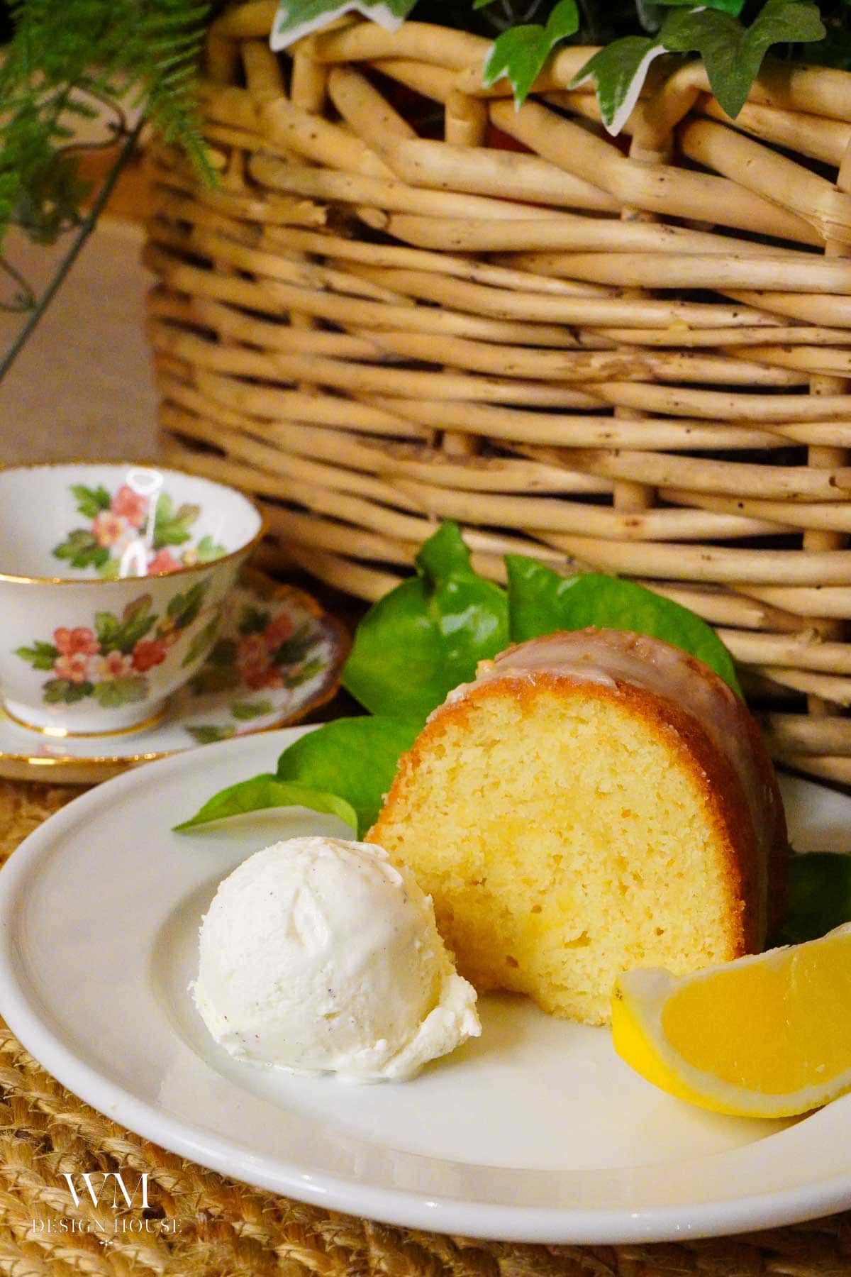 A slice of lemon curd Bundt cake with icing is served on a white plate alongside a scoop of vanilla ice cream, a lemon wedge, and green leaves. The cake is complemented by a slice of tangy Meyer Lemon. A floral teacup and a woven basket are in the background.