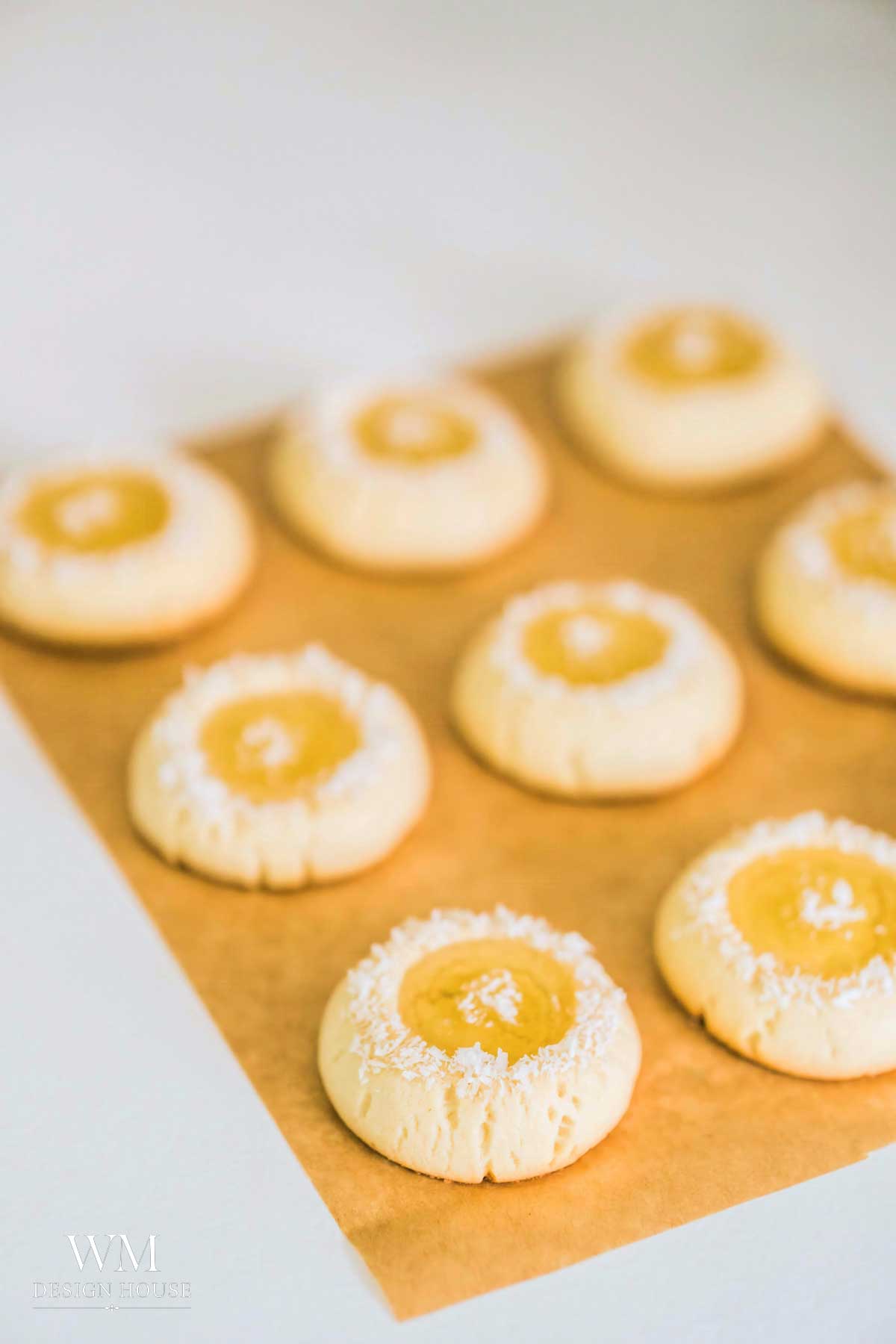 Eight round thumbprint cookies with bright lemon curd centers and white coconut sprinkles are arranged on a sheet of brown parchment paper against a light background.