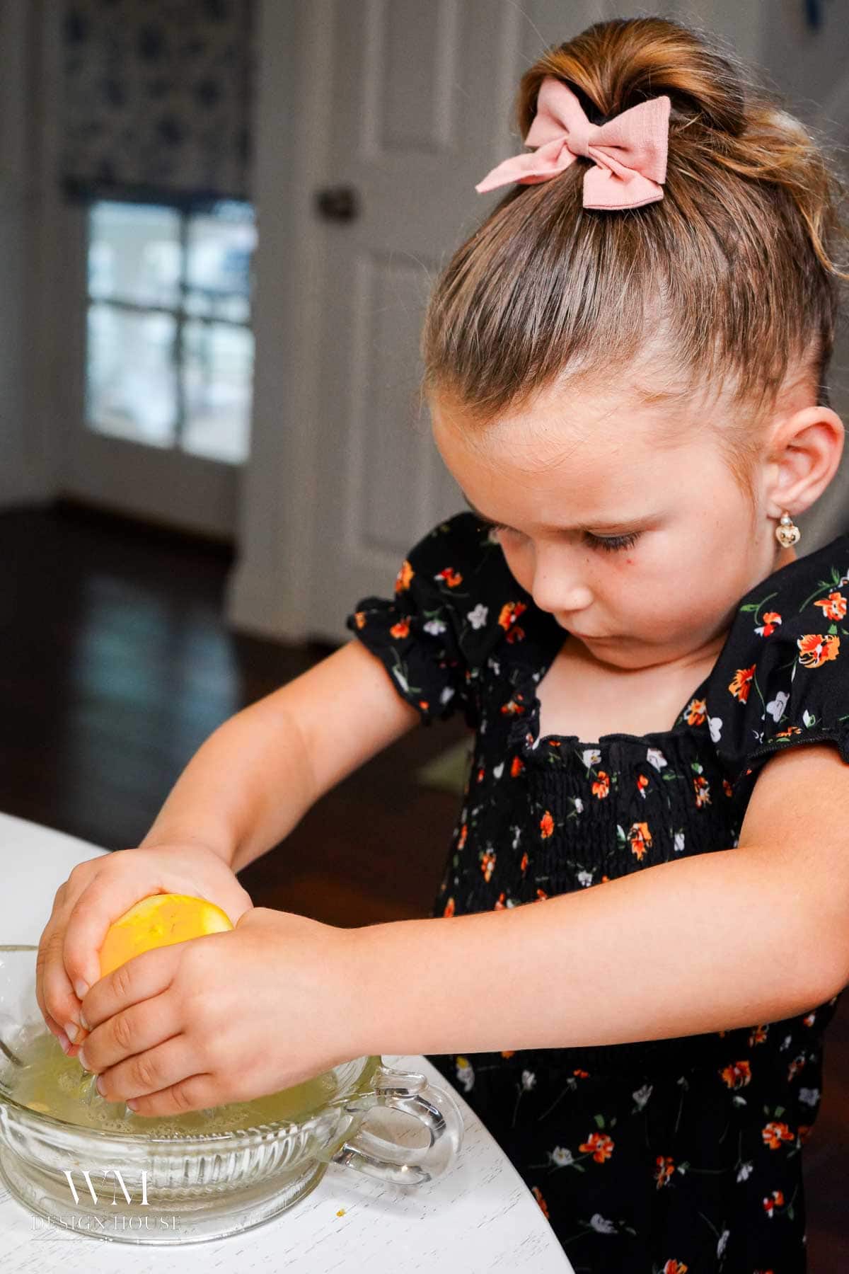 A young girl with a pink bow squeezes a lemon using a glass juicer in a kitchen, concentrating intently on her task.