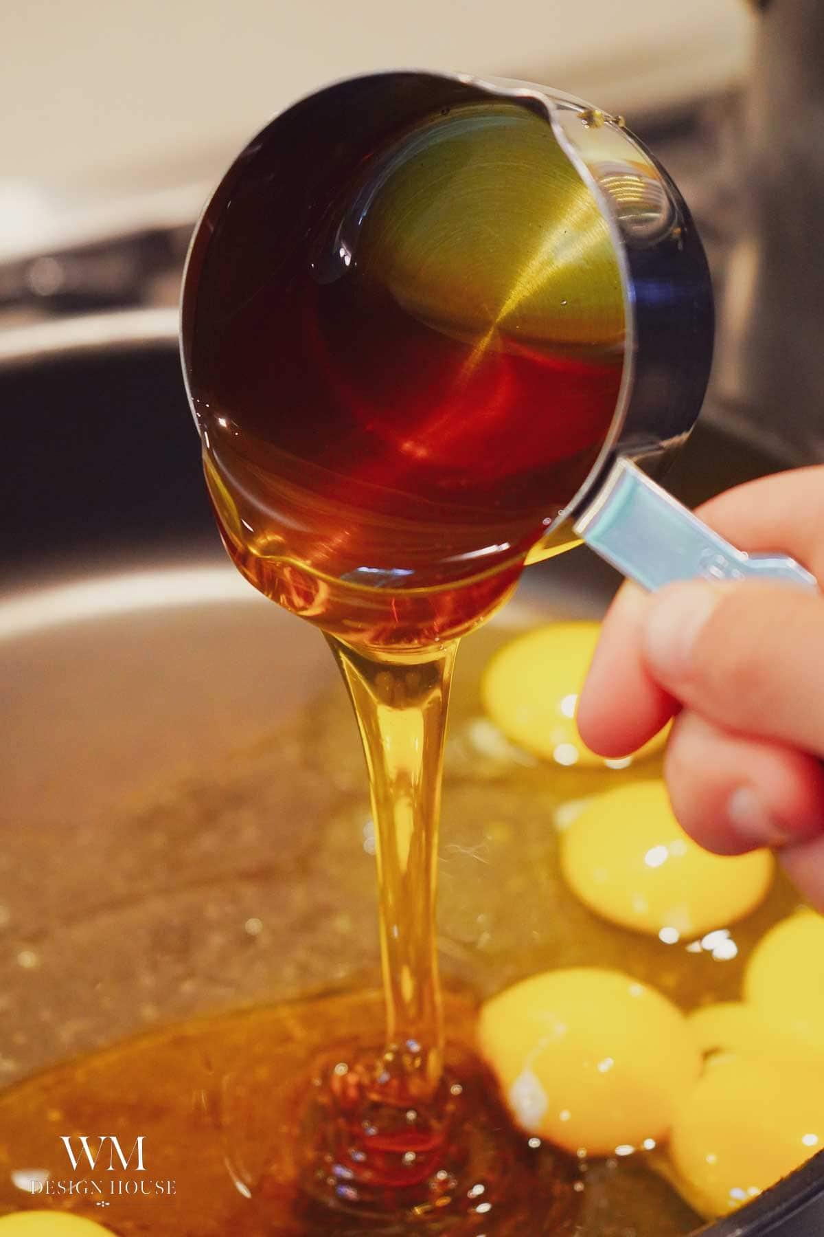 A hand pours a measuring cup of honey or syrup into a bowl containing several raw egg yolks. The liquid is golden and thick, cascading smoothly from the cup.
