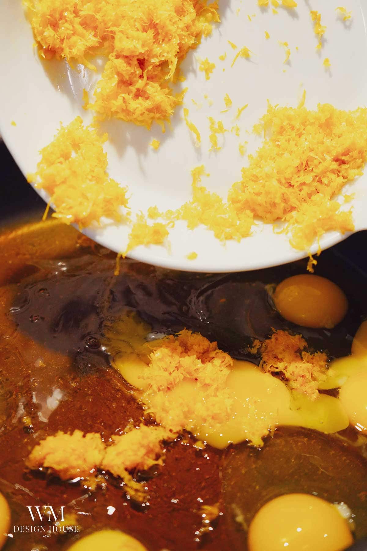 A white plate with orange zest is being poured into a bowl containing eggs and a dark liquid, possibly for baking or cooking.