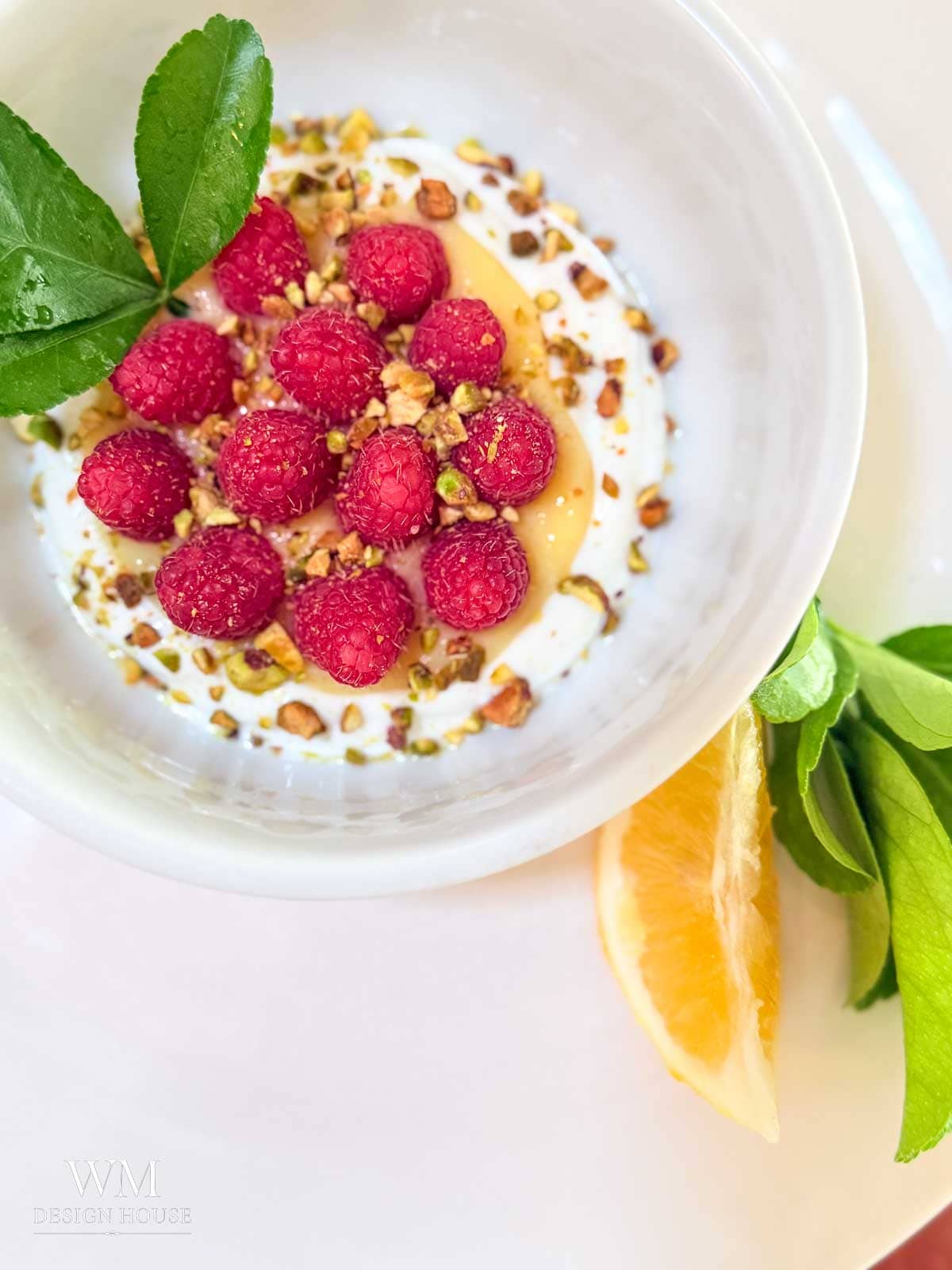 A white bowl with greek yogurt, lemon curd, fresh raspberries, chopped pistachios, honey, and green leaves is placed on a plate with a lemon wedge and more green leaves on the side. A drizzle of Salted Honey adds a unique touch of sweetness to this refreshing presentation.