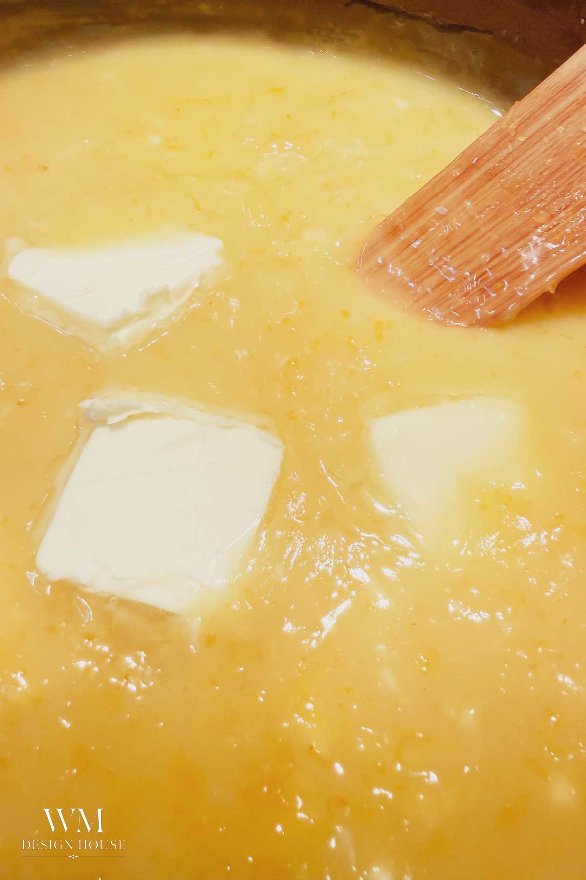 A close-up of a creamy yellow mixture with three chunks of butter and a wooden spatula stirring it.