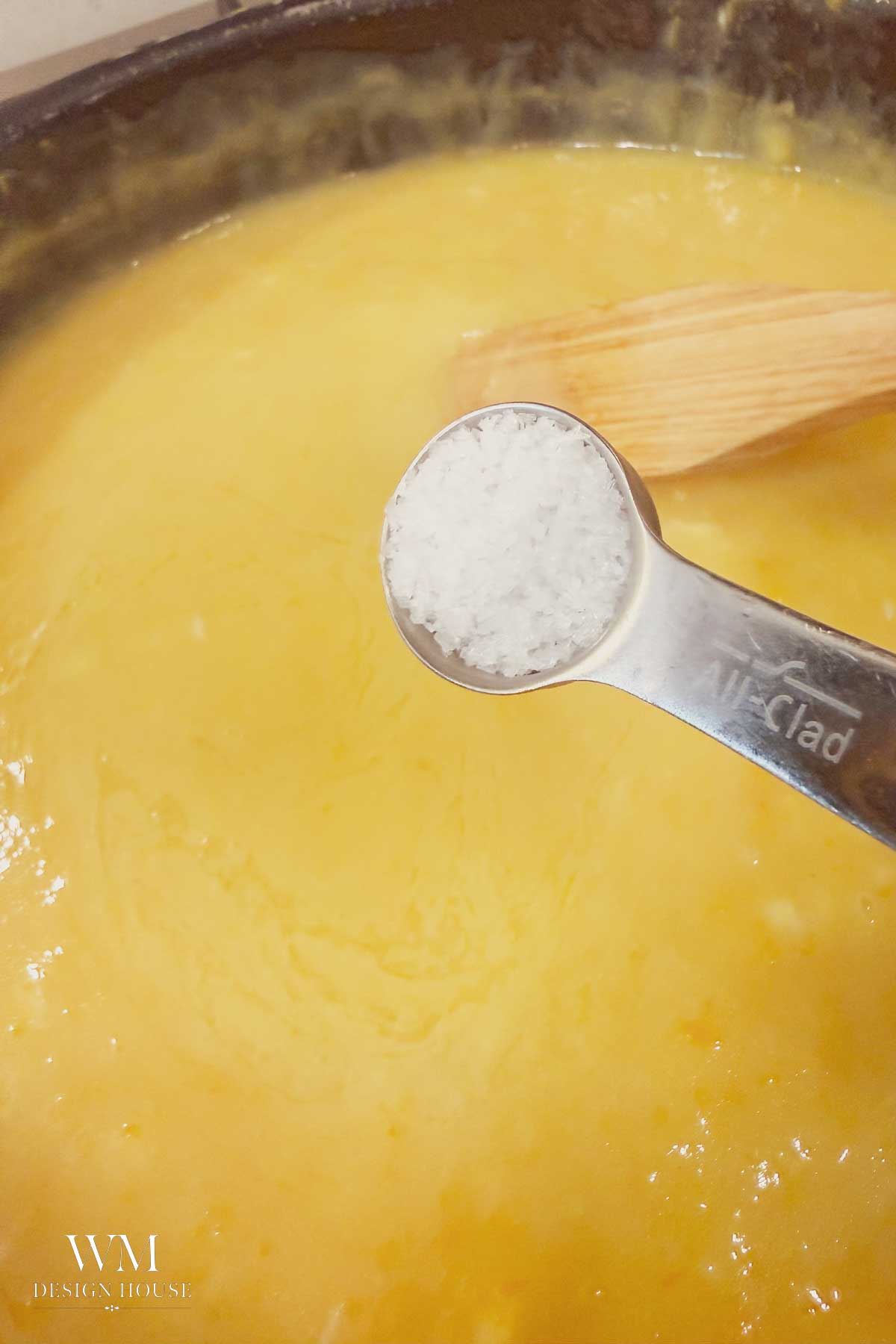A metal measuring spoon holding flaky salt is held above a pot of yellow sauce. A wooden spatula rests in the sauce.