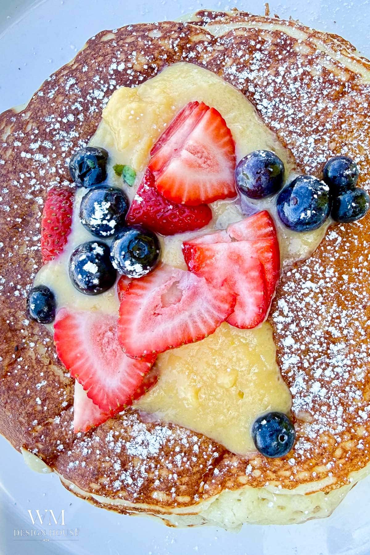 A close-up of a pancake topped with Meyer Lemon Curd, sliced strawberries, whole blueberries, and a dusting of powdered sugar.