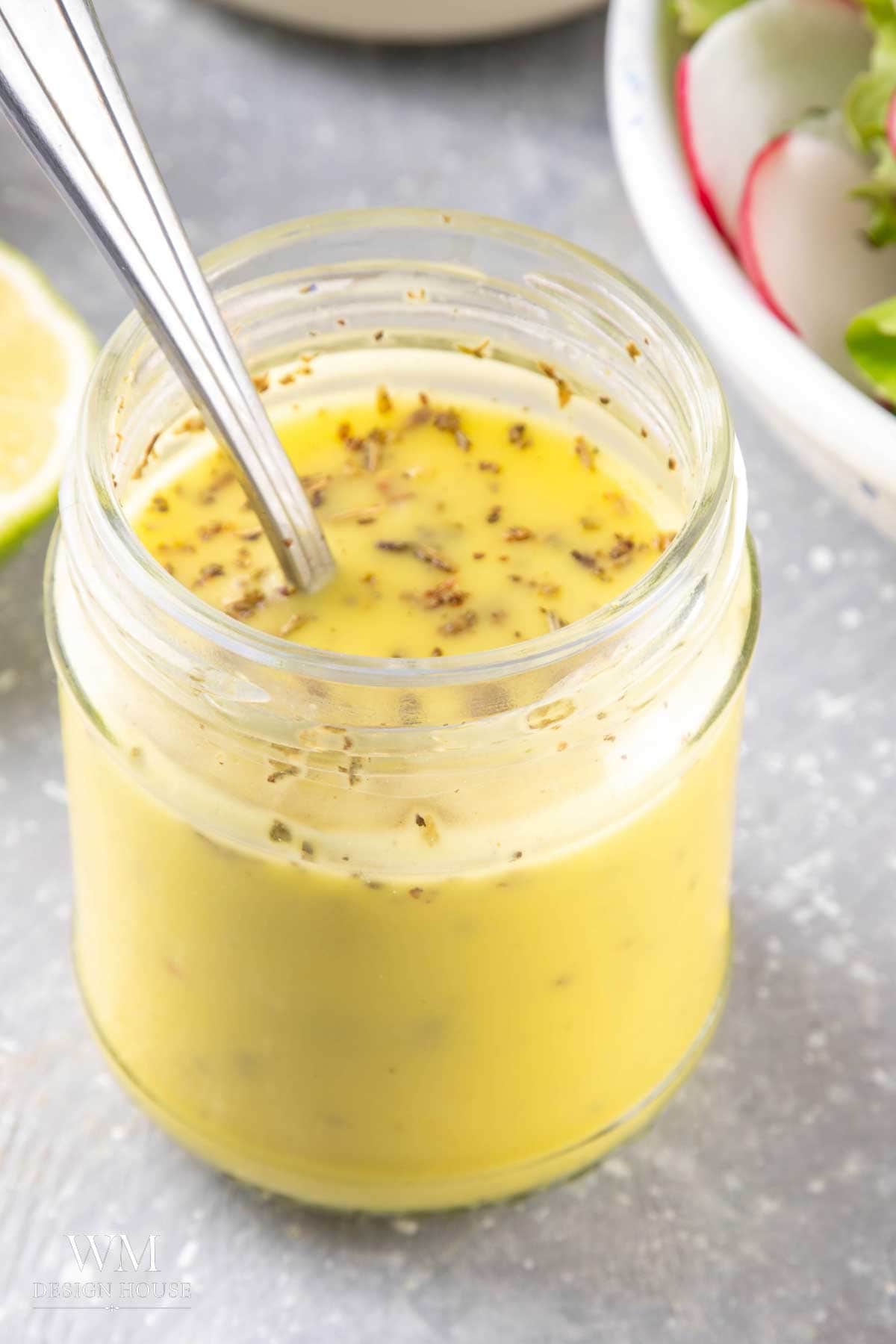 A glass jar filled with lemon curd salad dressing sprinkled with herbs, with a metal spoon inside; a bowl of salad and a lime wedge are blurred in the background.