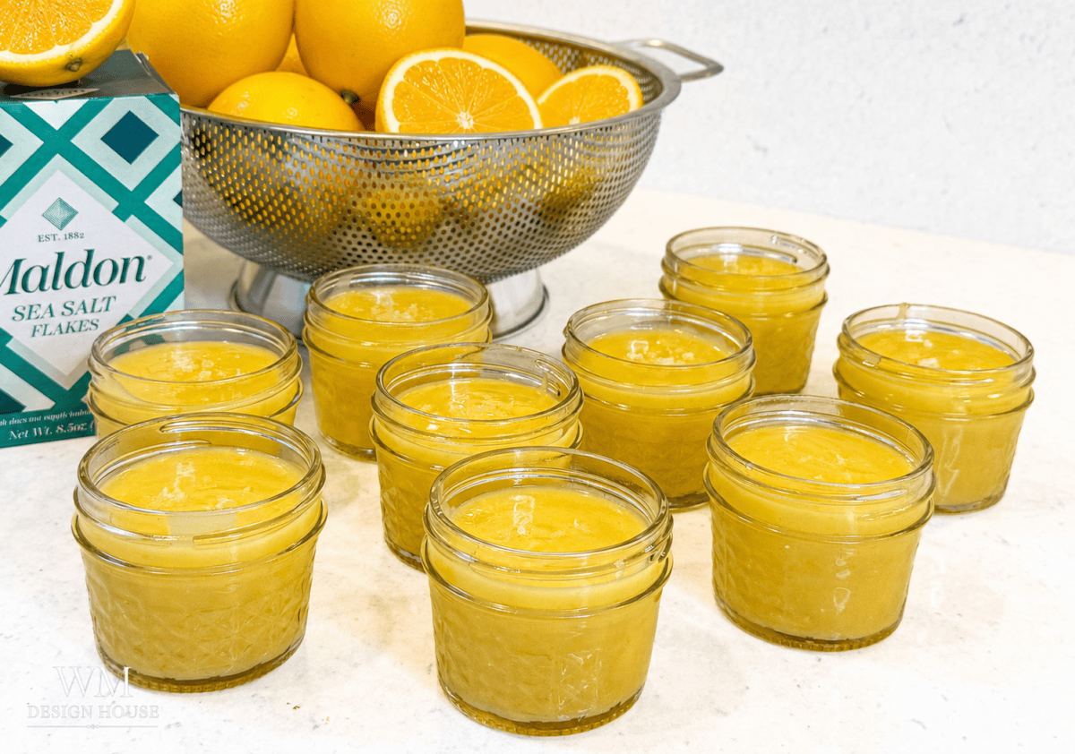 Eight small glass jars filled with vibrant Meyer Lemon Curd are arranged on a counter, with a colander of whole oranges and a box of Maldon sea salt flakes in the background.