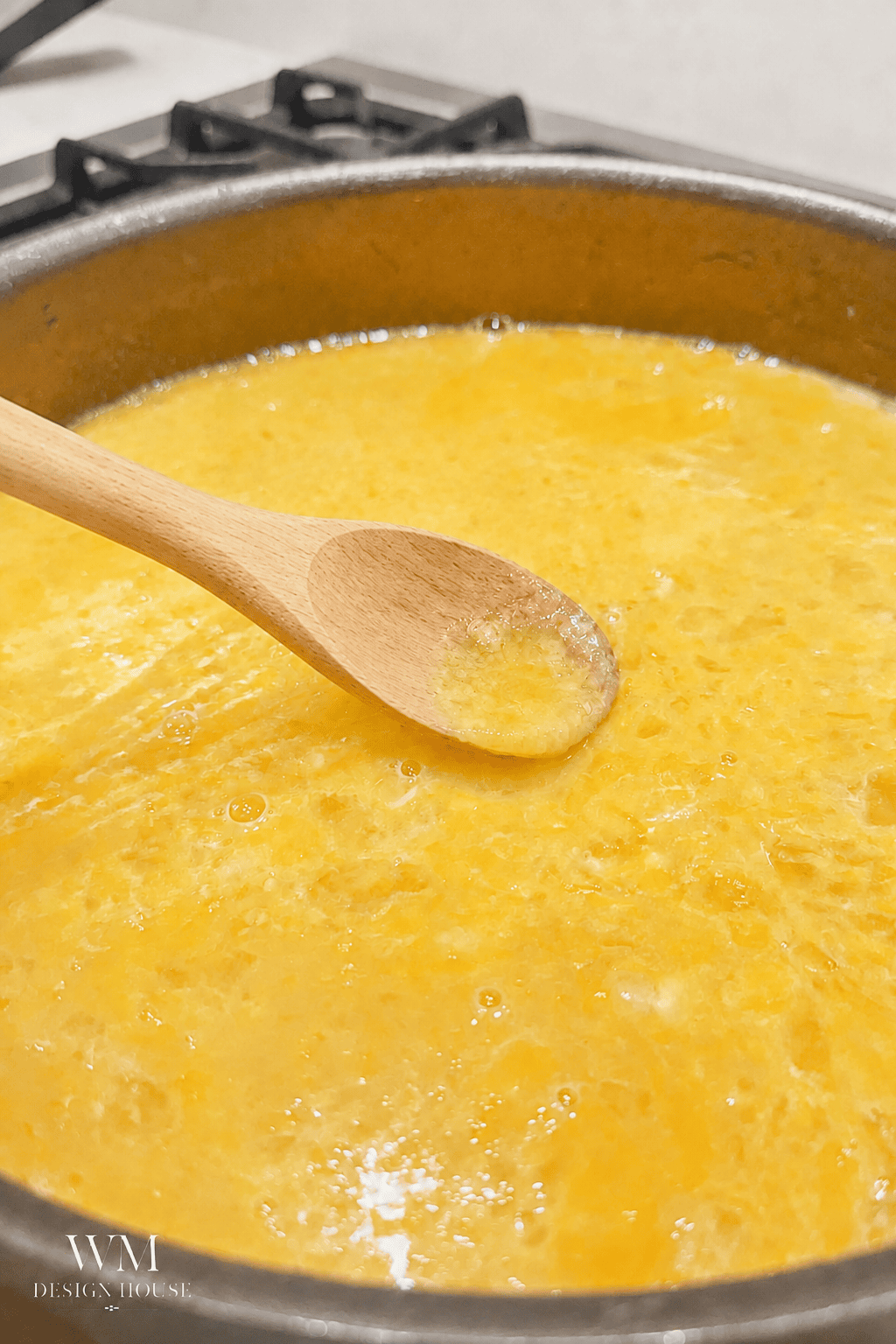 A close-up of scrambled eggs cooking in a pan, with a wooden spoon stirring them. A hint of Meyer Lemon Curd sits nearby, ready to add a bright finish. The stove burner is partially visible in the background.