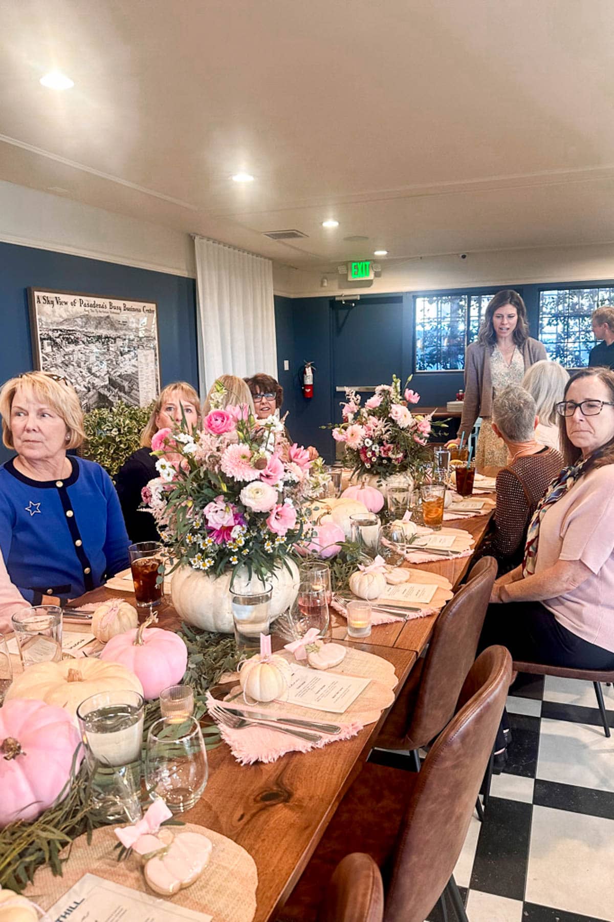 A group of women sit around a wooden table decorated with pink pumpkins and floral centerpieces, enjoying drinks in a cozy, well-lit room with checkered floors.