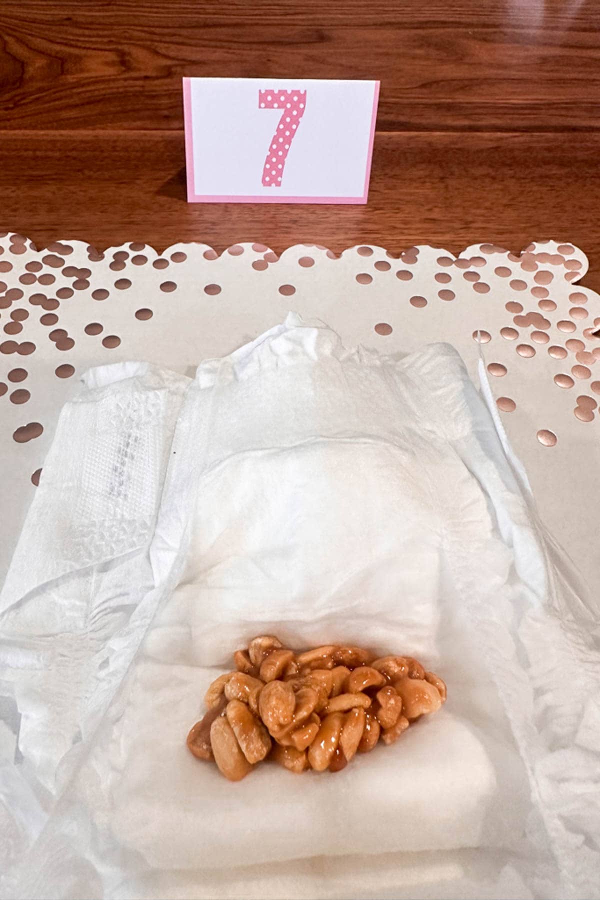 A diaper with what looks like caramel-covered peanuts sits on a table. Behind it is a pink and white card with the number 7. Confetti decorates the surface.