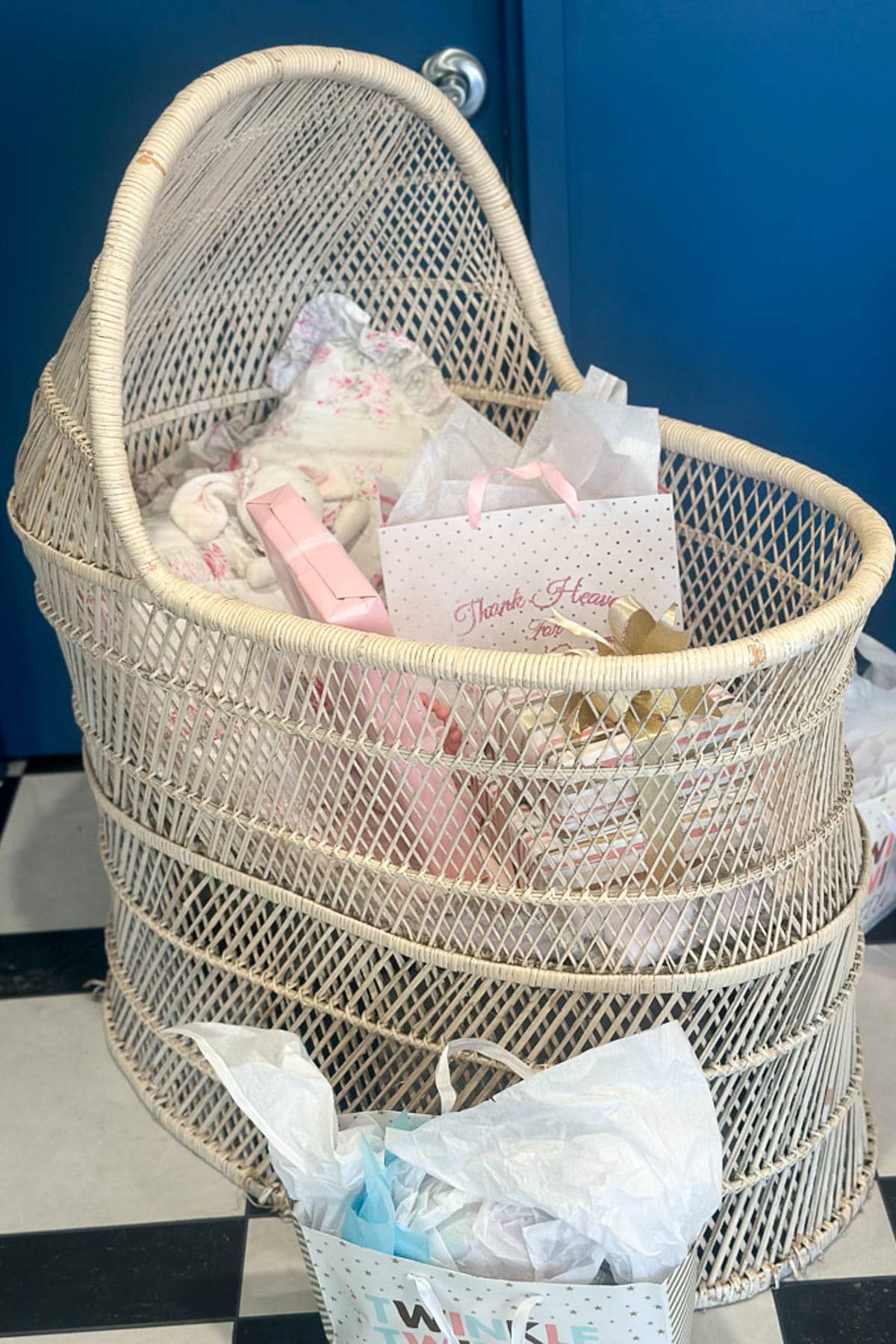 A woven bassinet filled with pastel-colored gift bags and tissue paper sits on a black and white tiled floor near a blue wall.