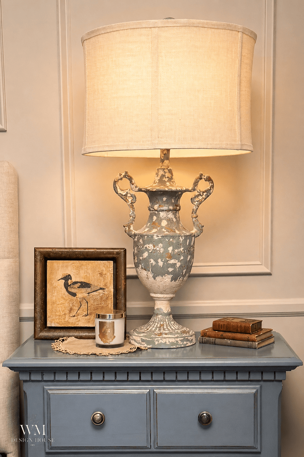 A vintage lamp with a wide shade sits on a blue nightstand alongside a framed bird picture, a candle, and two stacked books against a neutral paneled wall—perfect inspiration for cozy bedroom ideas and an inviting seasonal refresh.