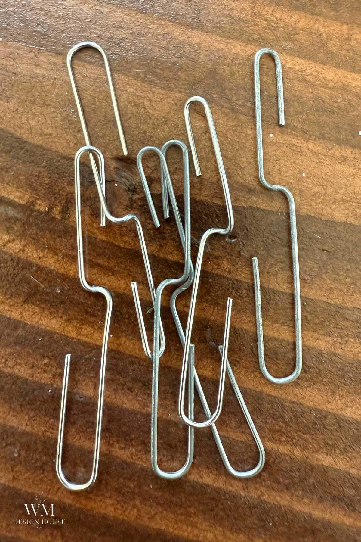 Several silver S-shaped paperclips are scattered on a wooden surface, reminiscent of the careful arrangement needed when learning how to dry dahlias. The grain of the wood is visible, and the paperclips overlap each other in a loose pile.