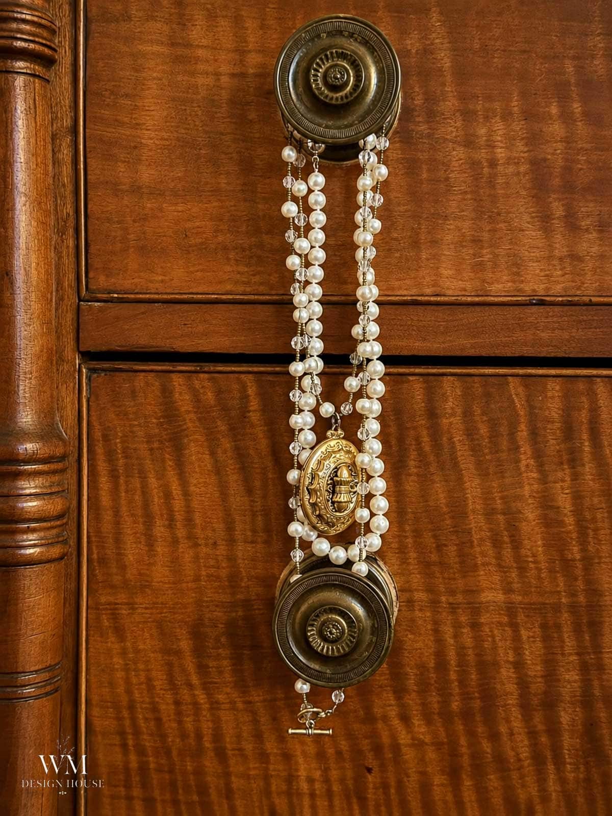 A string of pearls with a gold pendant is draped over two ornate brass drawer knobs on a wooden cabinet, adding a touch of cozy fall bedroom decor. The “WM Design House” logo appears in the bottom left corner.