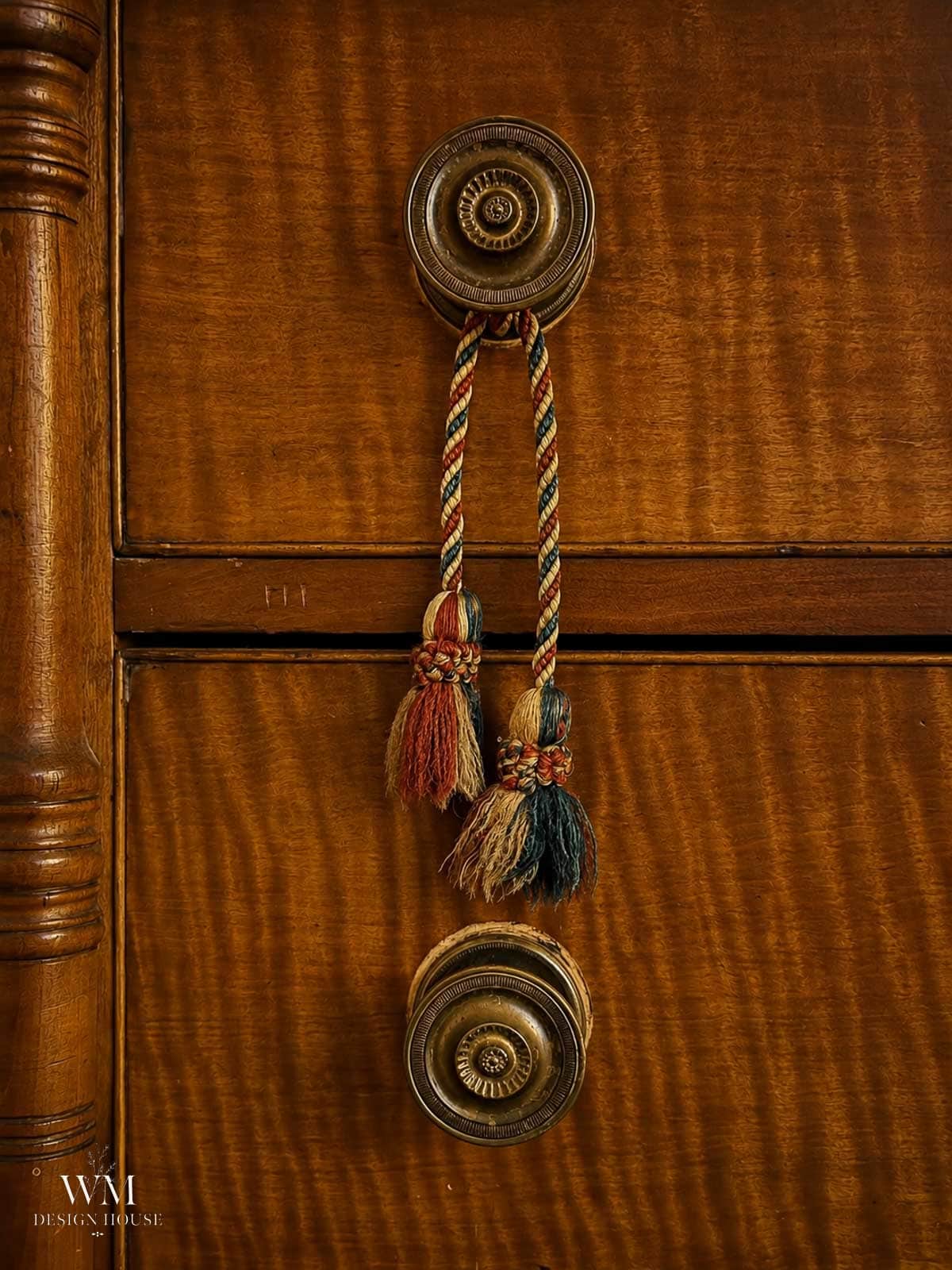 Two ornate brass drawer knobs on a wooden dresser are connected by a braided cord with red, green, and beige tassels hanging in the center—a charming accent that’s perfect for cozy fall bedroom decor. Subtle wood grain patterns are visible in the background, adding warmth and texture for a seasonal refresh.