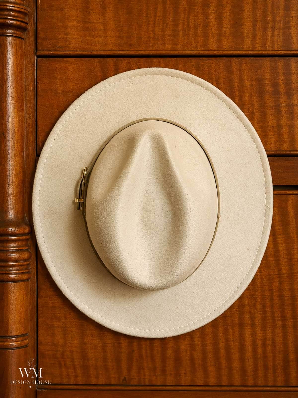 A beige felt hat with a wide brim hangs on a wooden wardrobe door, adding a touch of cozy fall decor to the space. The hat is centered in the image, and the wood has a warm brown finish. “WM DESIGN HOUSE” is written in the bottom left corner.