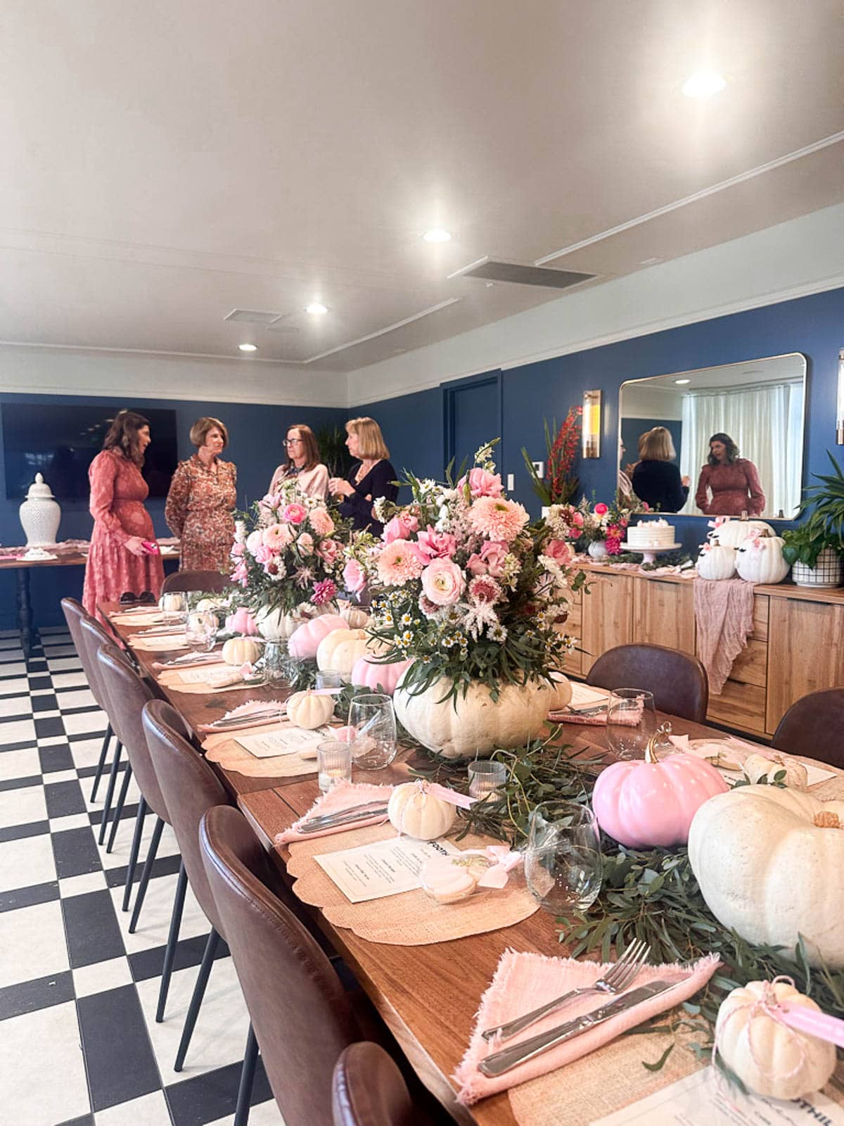 Elegant dining table with pink and white floral pumpkin centerpieces, place settings, and menus. Four women stand and chat near the end of the table in a stylish room with blue walls and checkered flooring.