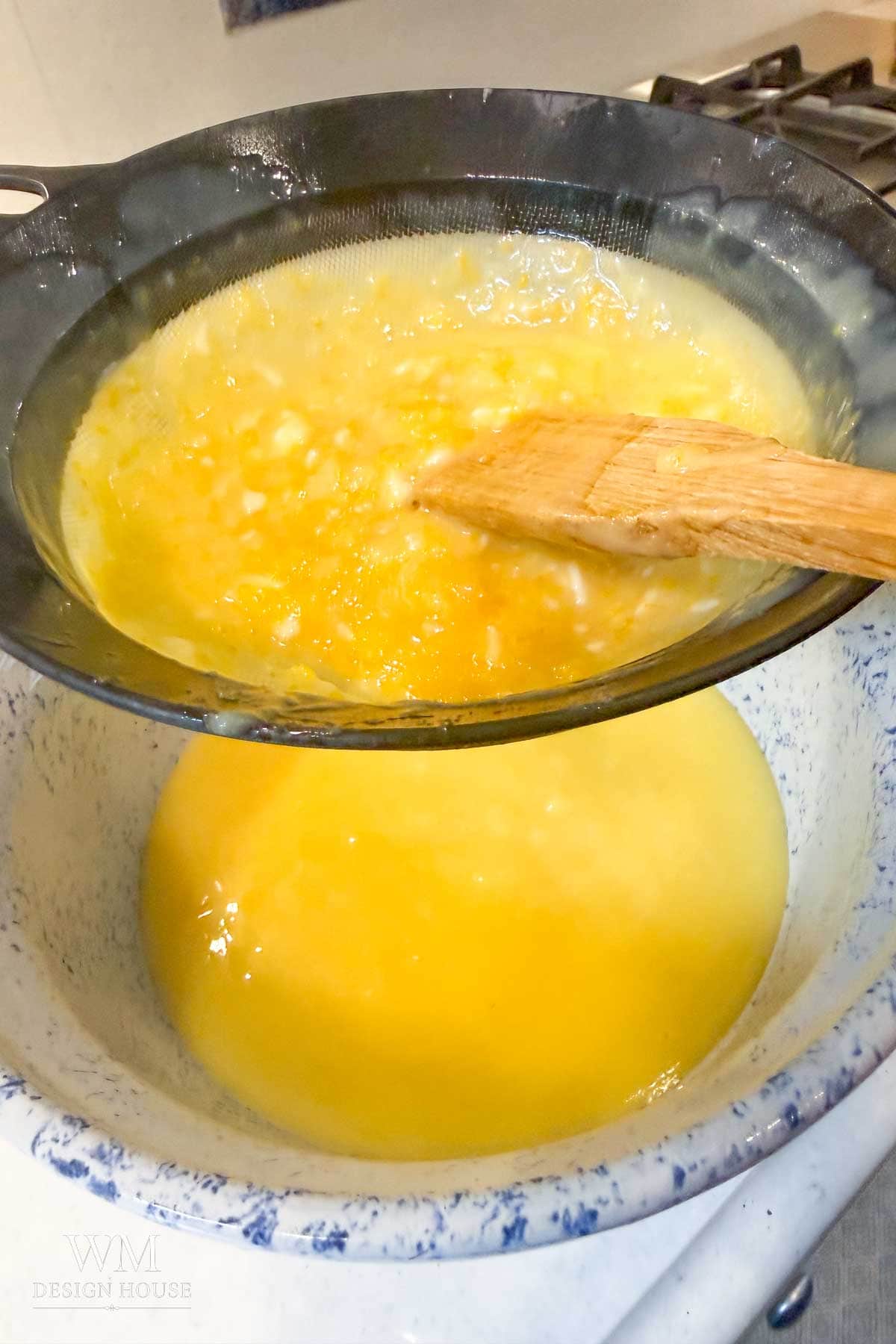 A wooden spoon stirs melted butter in a mesh strainer, separating milk solids from clarified butter over a speckled bowl on a stovetop, perfect for making silky Meyer Lemon Curd or salted honey infusions.