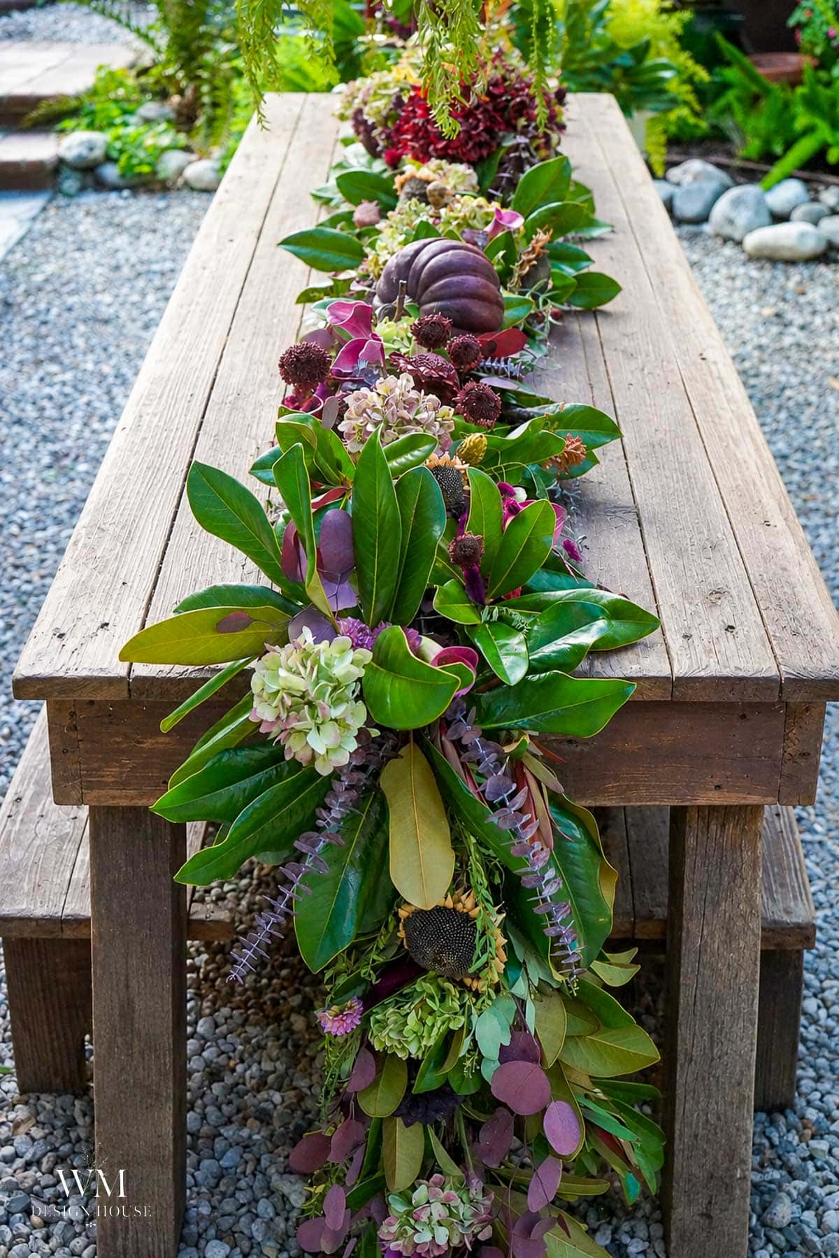 Styled greenery table runner with dried dahlias, antique hydrangeas, and a plum pumpkin down a bare wood table