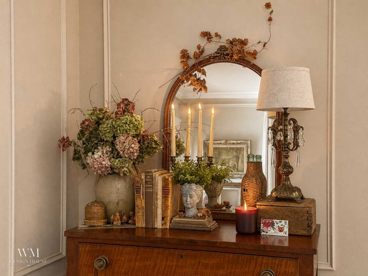 A wooden dresser with a mirror displays dried flowers, candles, vintage books, a bust planter, a lamp, a candle, and decorative items. Warm, cozy, vintage decor is reflected in the mirror.