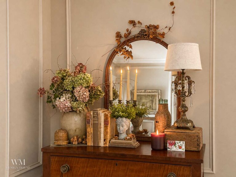 A wooden dresser with a mirror holds dried flowers, books, a bust planter, candles, a lamp, and decorative items, creating a cozy, vintage-inspired vignette—perfect for those seeking cozy bedroom ideas or planning a fall bedroom decor seasonal refresh.