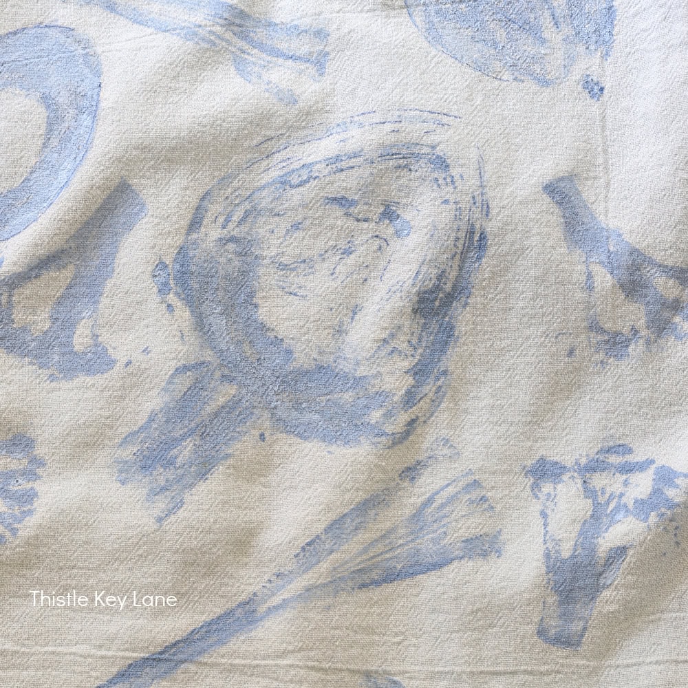 A white cloth features pale blue vegetable prints, including mushrooms and broccoli. The corner shows the text “Thistle Key Lane” in small white font.