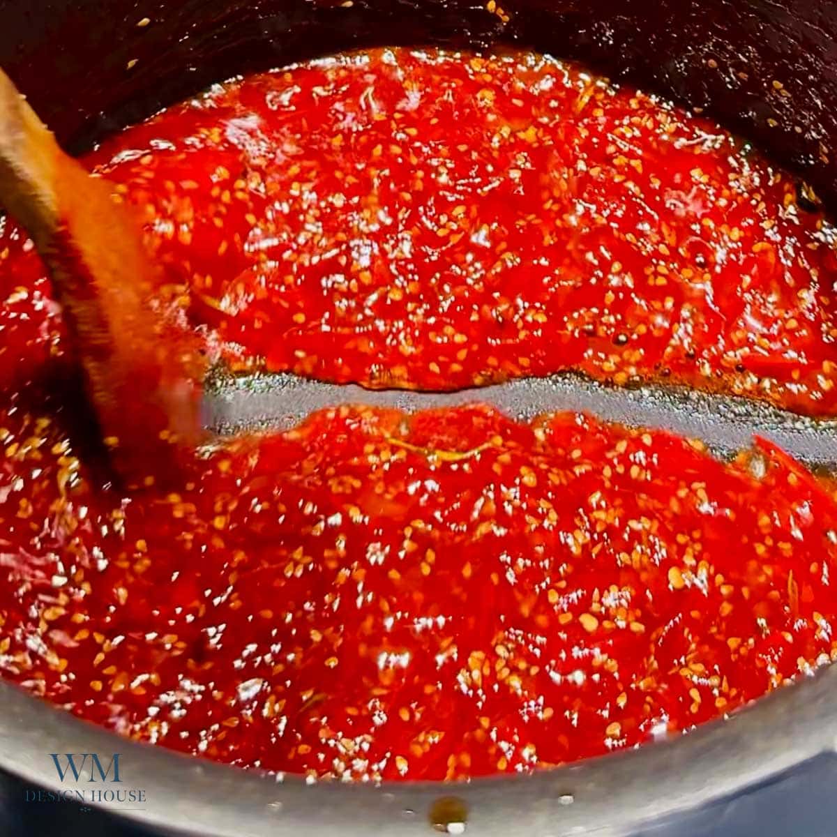 A pot of red tomato jam with visible seeds is being stirred with a wooden spoon. The sauce appears thick and chunky.