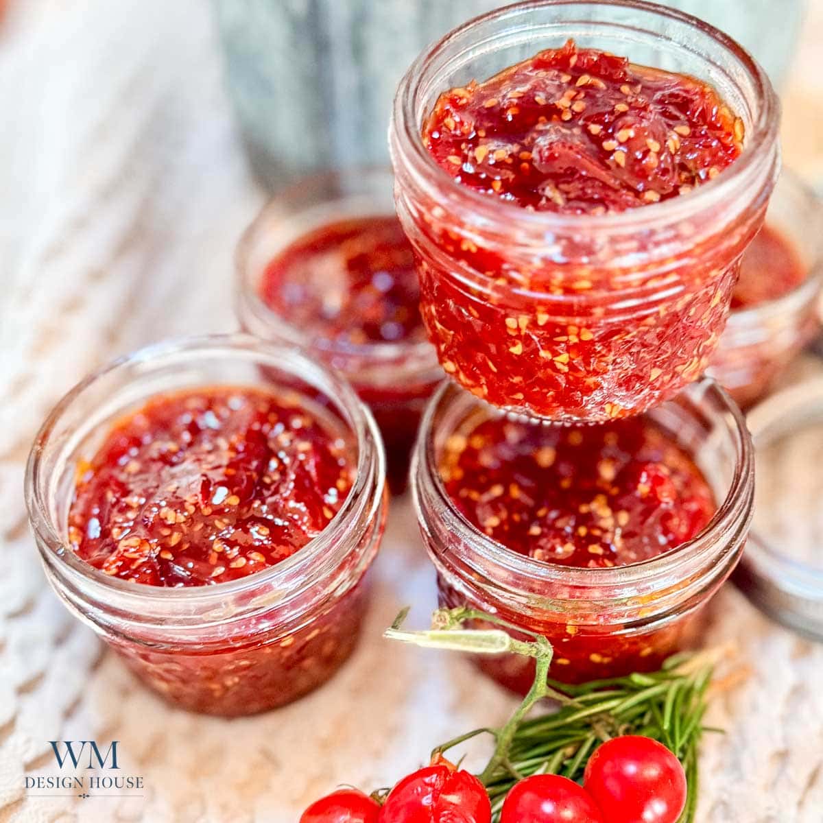 Four small glass jars filled with homemade raspberry or strawberry jam are arranged on a surface; a sprig of rosemary and red berries are nearby.