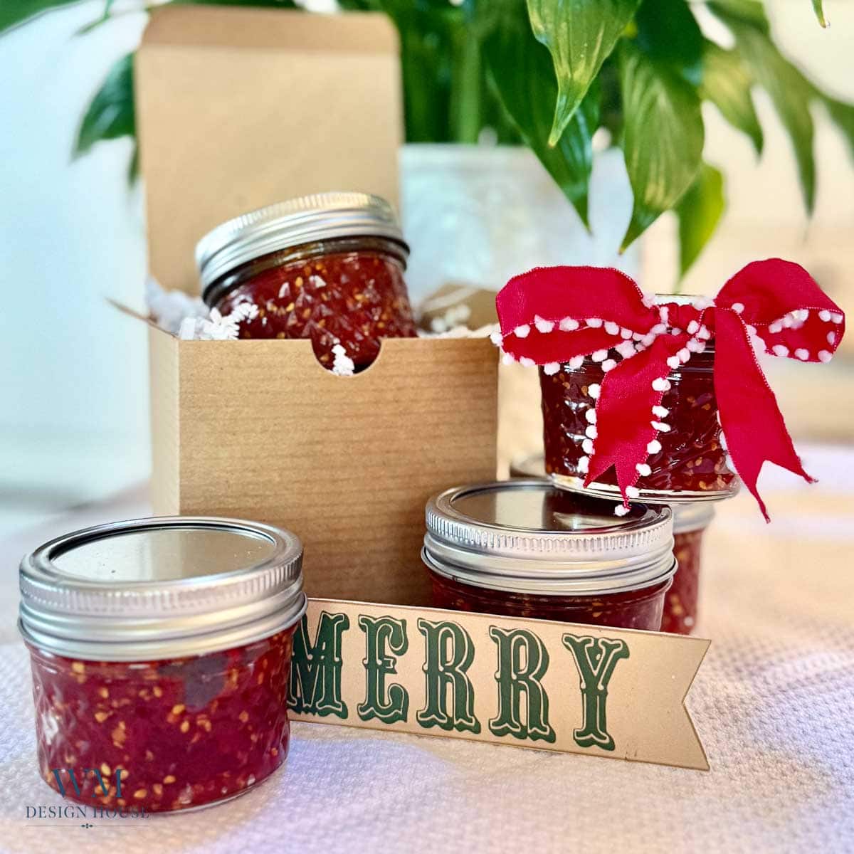 Four jars of homemade jam, one with a red ribbon, are displayed with a “MERRY” tag and a cardboard gift box. A green plant is in the background.