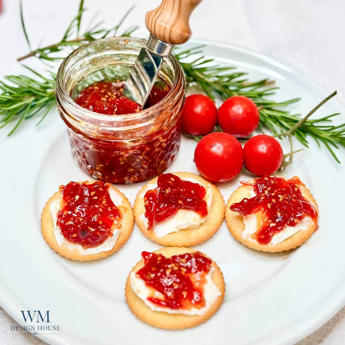 A plate with crackers topped with cheese and red jam, a jar of jam with a knife, sprigs of rosemary, and three red cherry tomatoes.