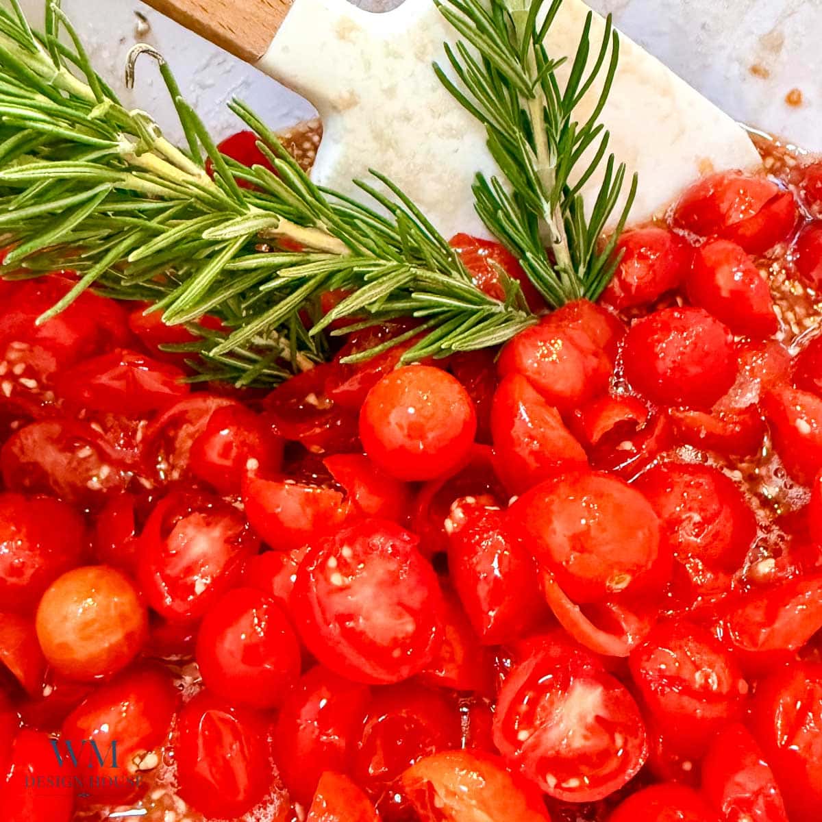 Chopped cherry tomatoes with sprigs of fresh rosemary on a white cutting board, with a white spatula partially visible in the background.