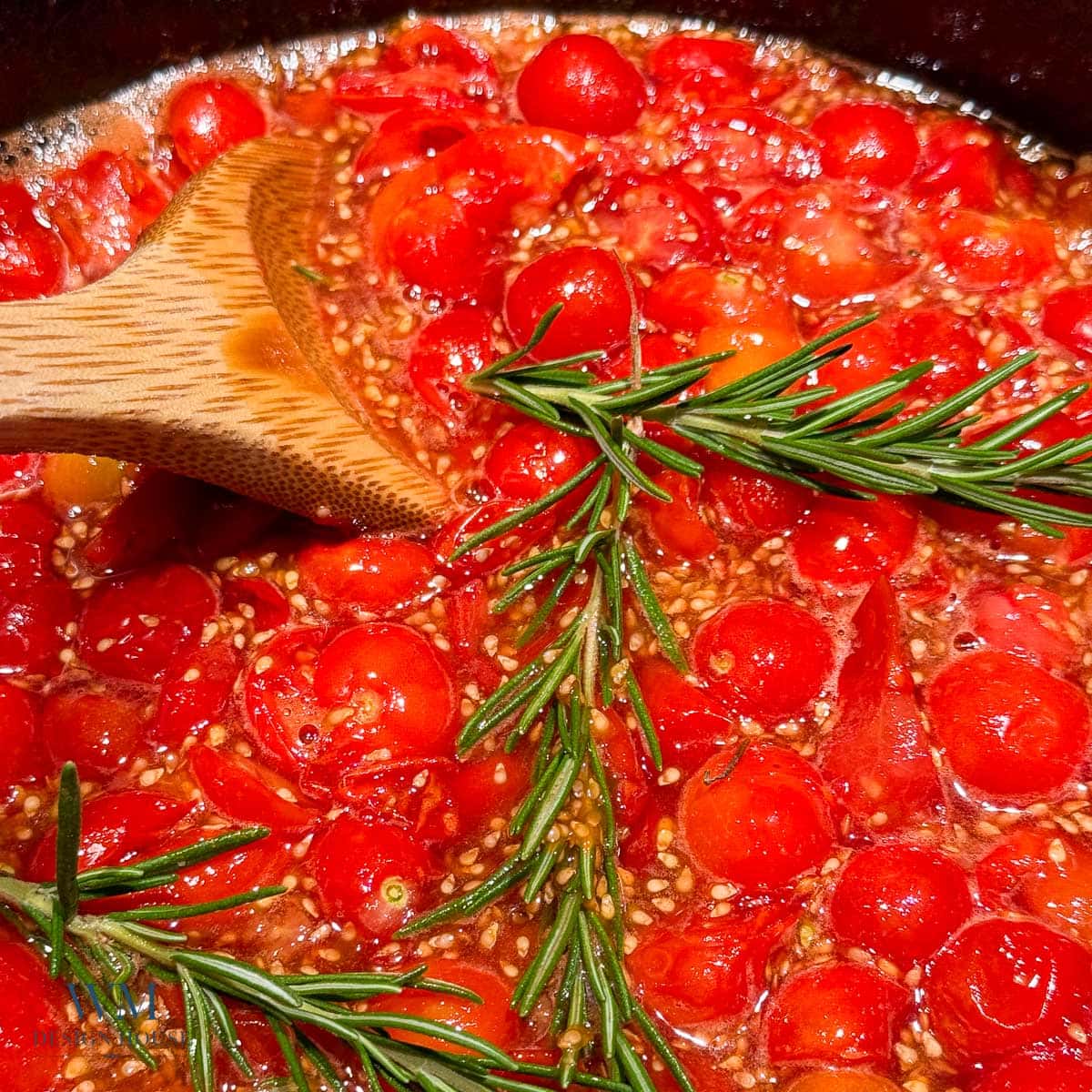 Chopped cherry tomatoes simmer in a pan with sprigs of fresh rosemary and a wooden spoon stirring the mixture.