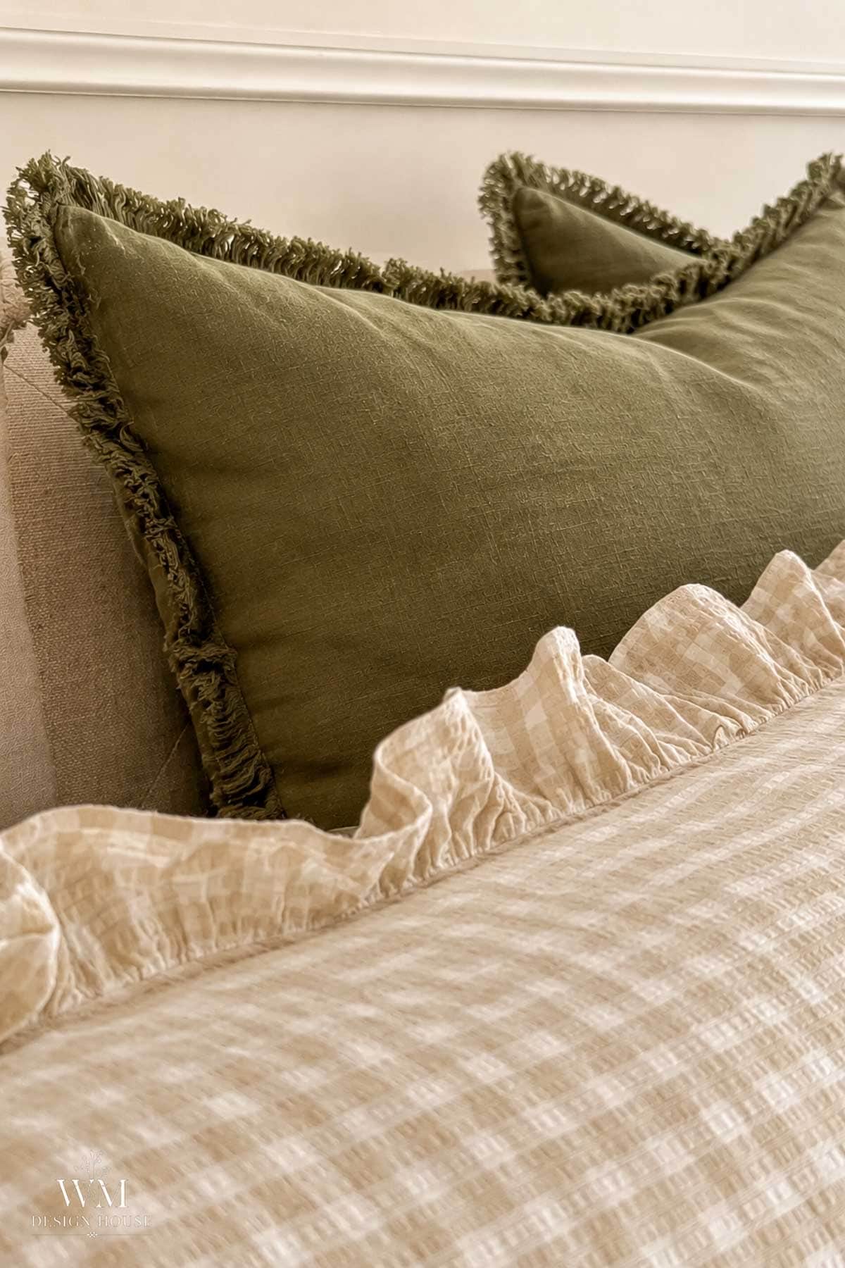 A close-up of a bed with a beige gingham blanket, a ruffled edge, and an olive green pillow with fringed trim, against a neutral background.