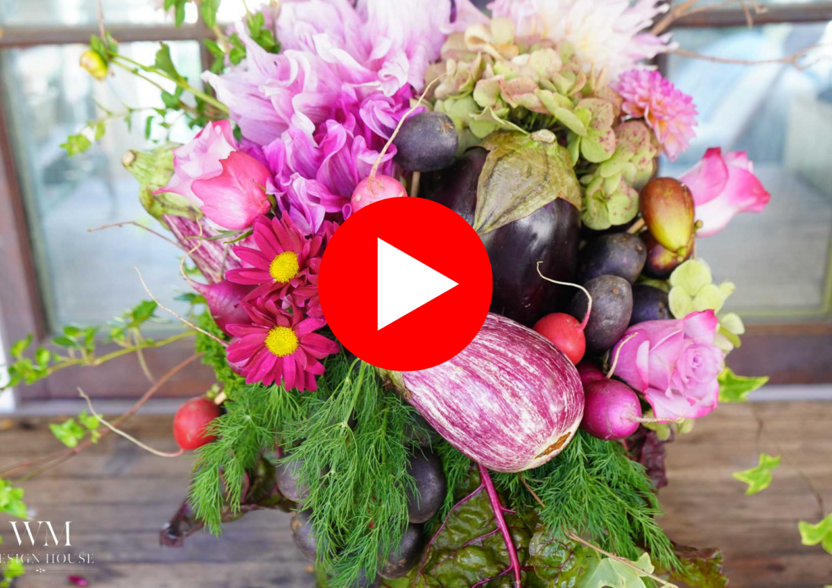 A vibrant vegetable-centerpiece bouquet mixing pink flowers and various vegetables, including eggplant and radishes, is arranged in front of a window. A large red play button icon is centered on the image.