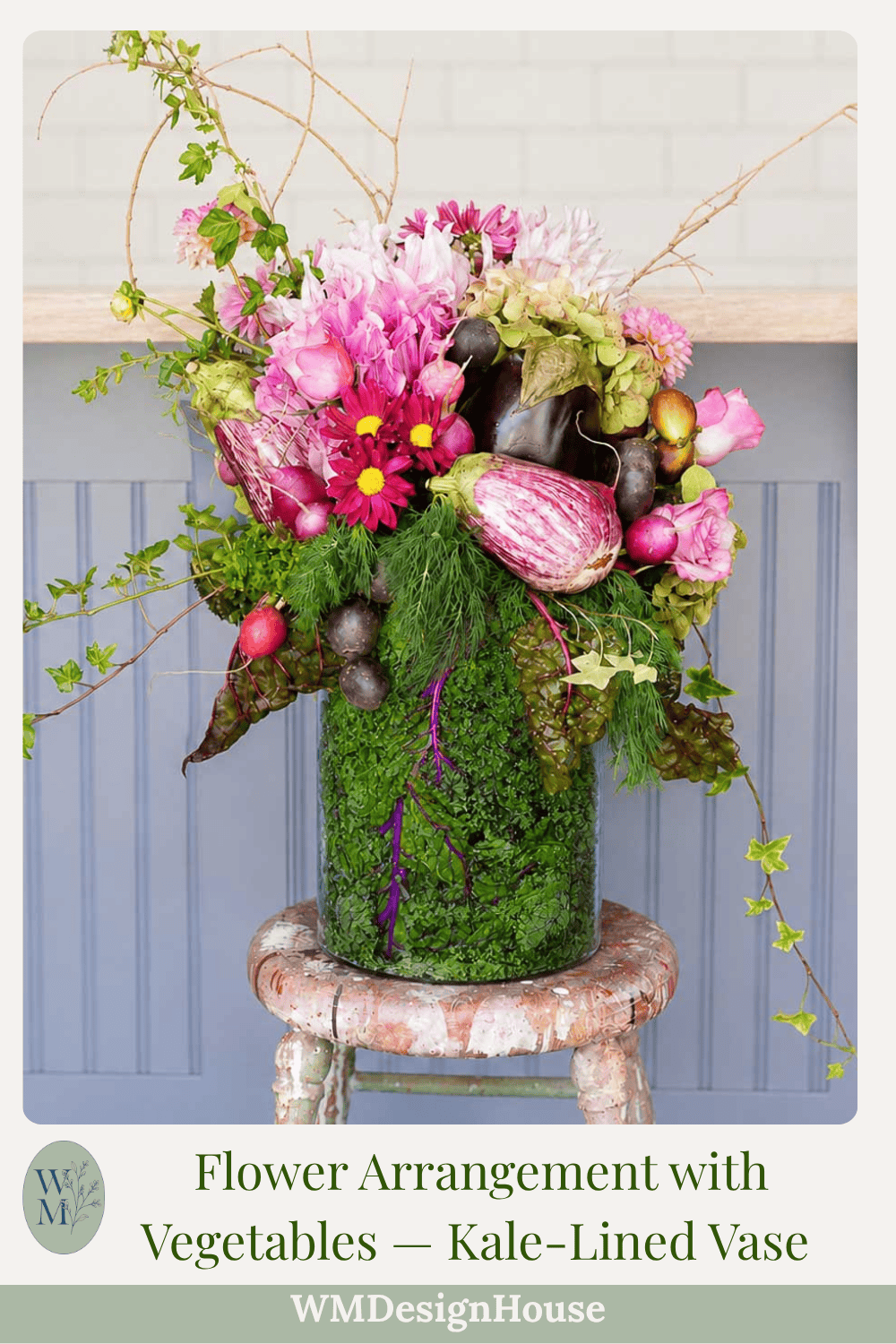 A vibrant vegetable-centerpiece features a unique flower arrangement mixed with vegetables in a clear vase lined with kale, sitting on a rustic stool against a light gray background. Pink flowers and assorted produce like eggplants and radishes are visible.