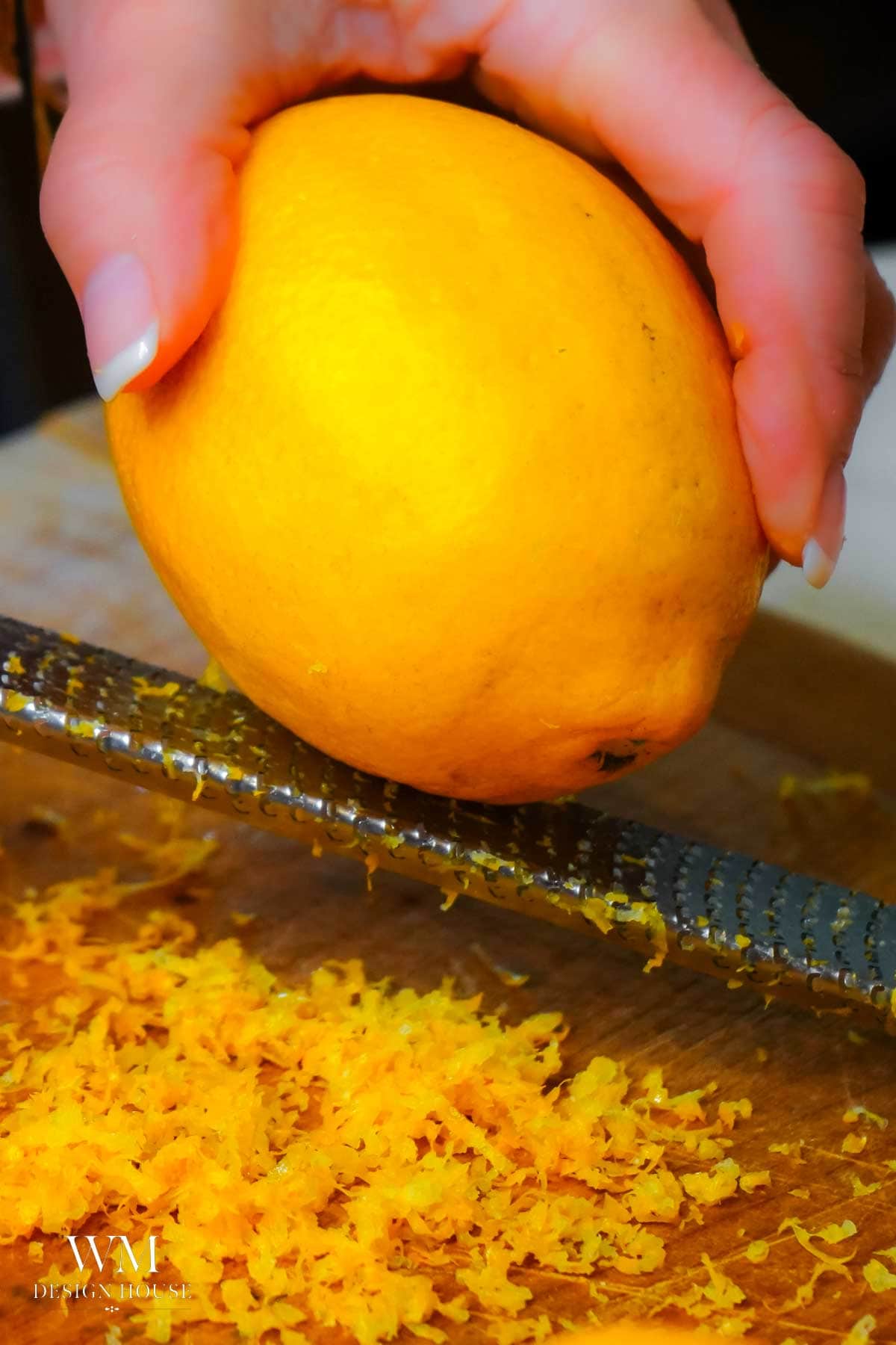 A hand grates the zest from a lemon onto a wooden cutting board, creating a pile of fine yellow zest below the grater—the perfect aromatic addition for recipes like Lemon Curd or Salted Honey desserts.
