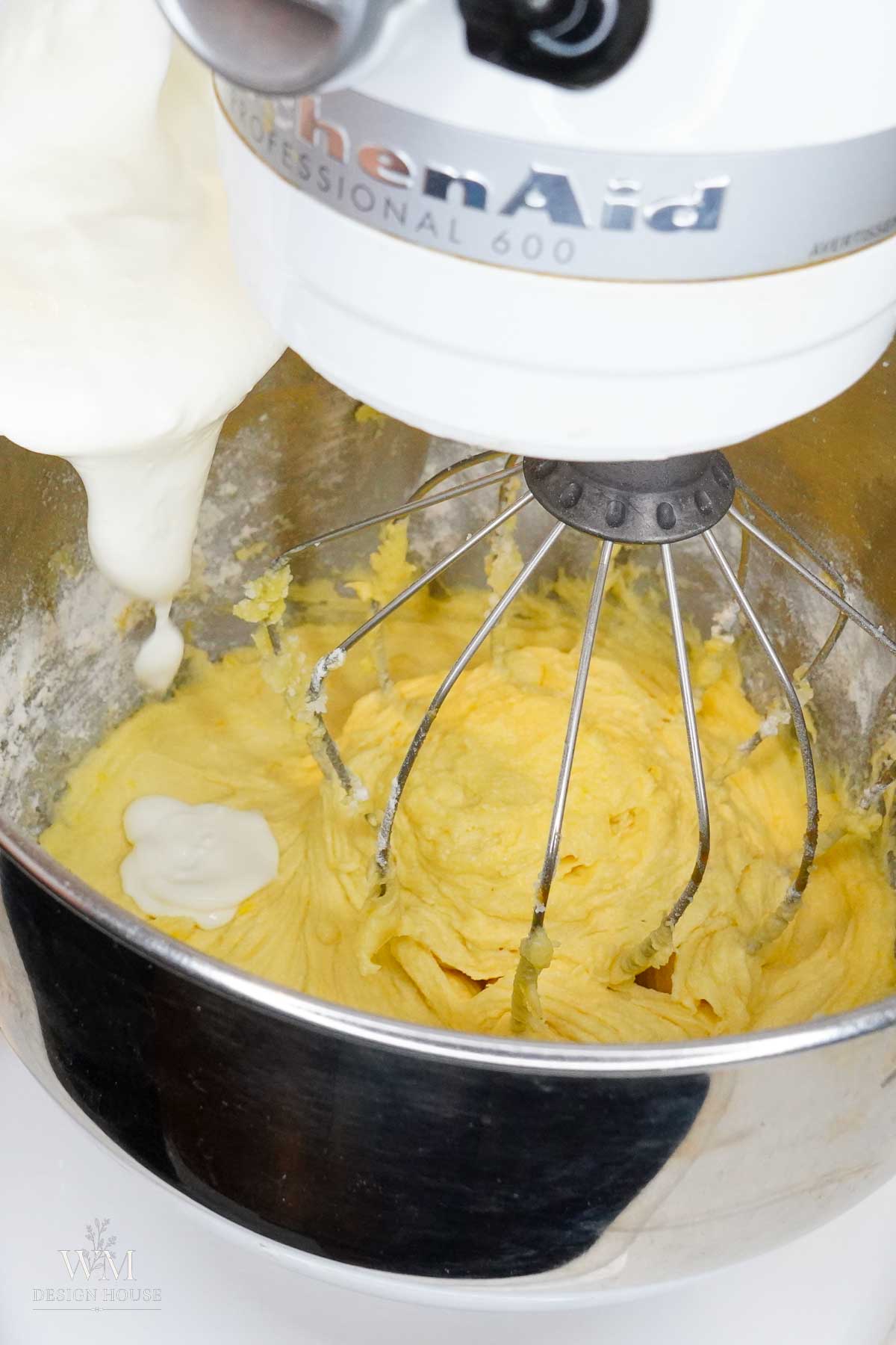 A stand mixer with yellow batter in the bowl and a whisk attachment is being used. A thick, white liquid is being poured into the bowl as the mixer prepares the mixture.