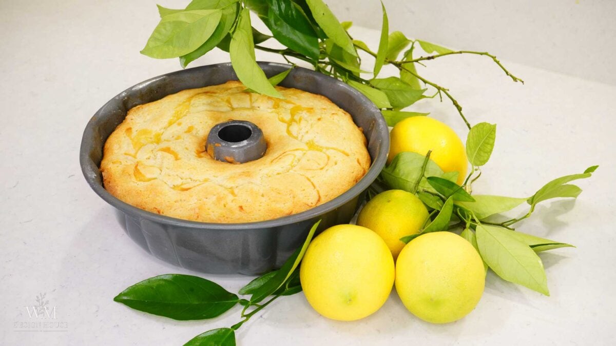 A freshly baked Bundt cake in a pan sits next to whole lemons and green lemon leaves on a white surface.