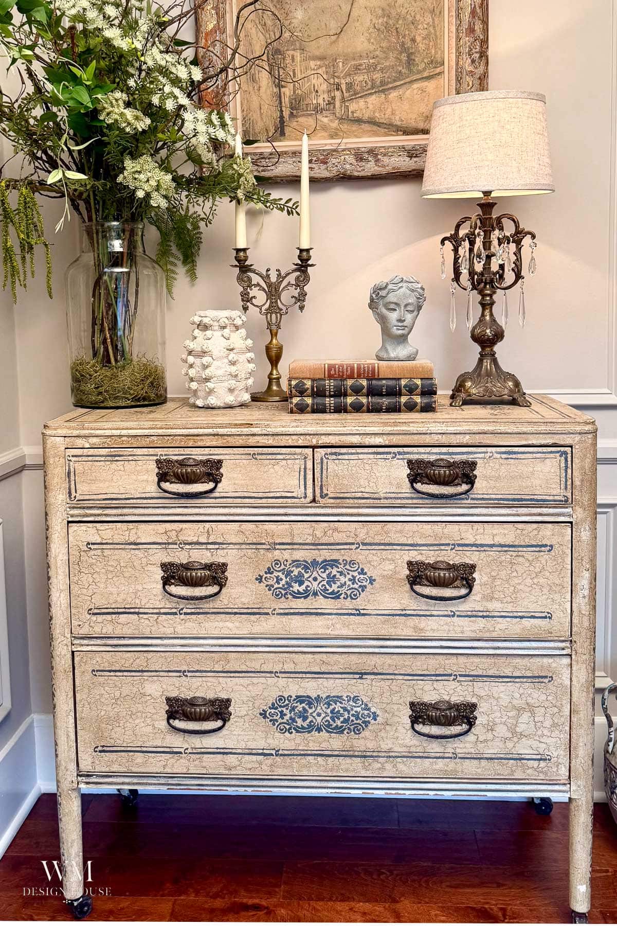 Ornate vintage dresser with decorative handles, topped with books, a bust, flowers, candlesticks, and a lamp. Framed art hangs above; the scene is classic and elegant—perfect inspiration for those seeking timeless fall bedroom decor ideas.