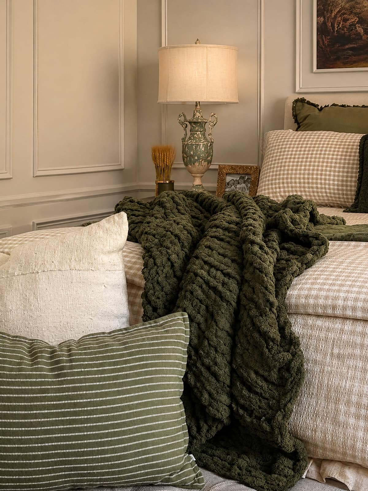 A cozy bedroom with beige gingham bedding, an olive green throw, striped and solid pillows, a classic nightstand with a lamp, a decorative vase, and framed photos, all in warm, neutral tones.