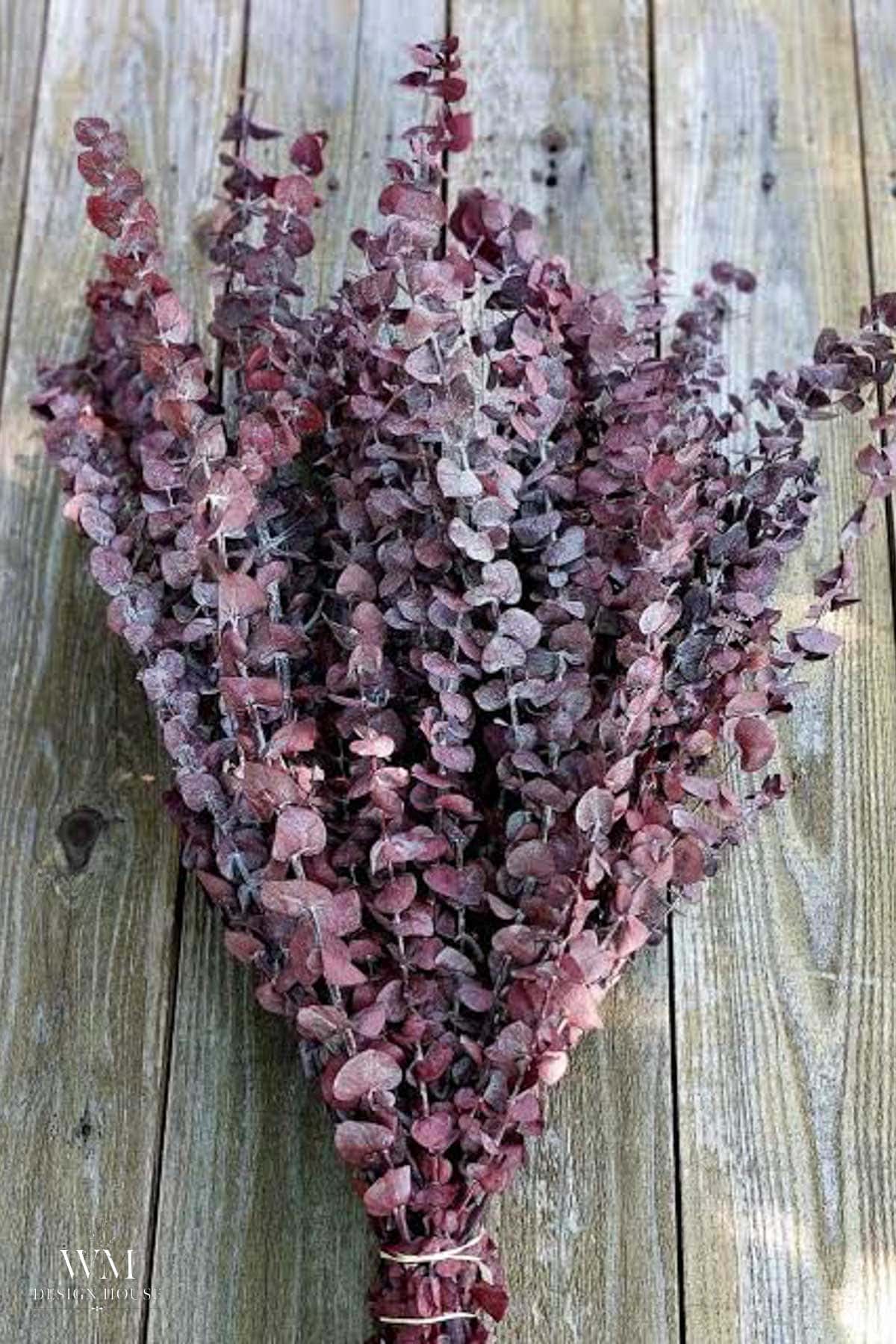 A bundle of dried purple eucalyptus branches lies on a rustic wooden surface, their small round leaves and varying shades of purple and dusty lavender tones creating the perfect accent for a greenery table runner.