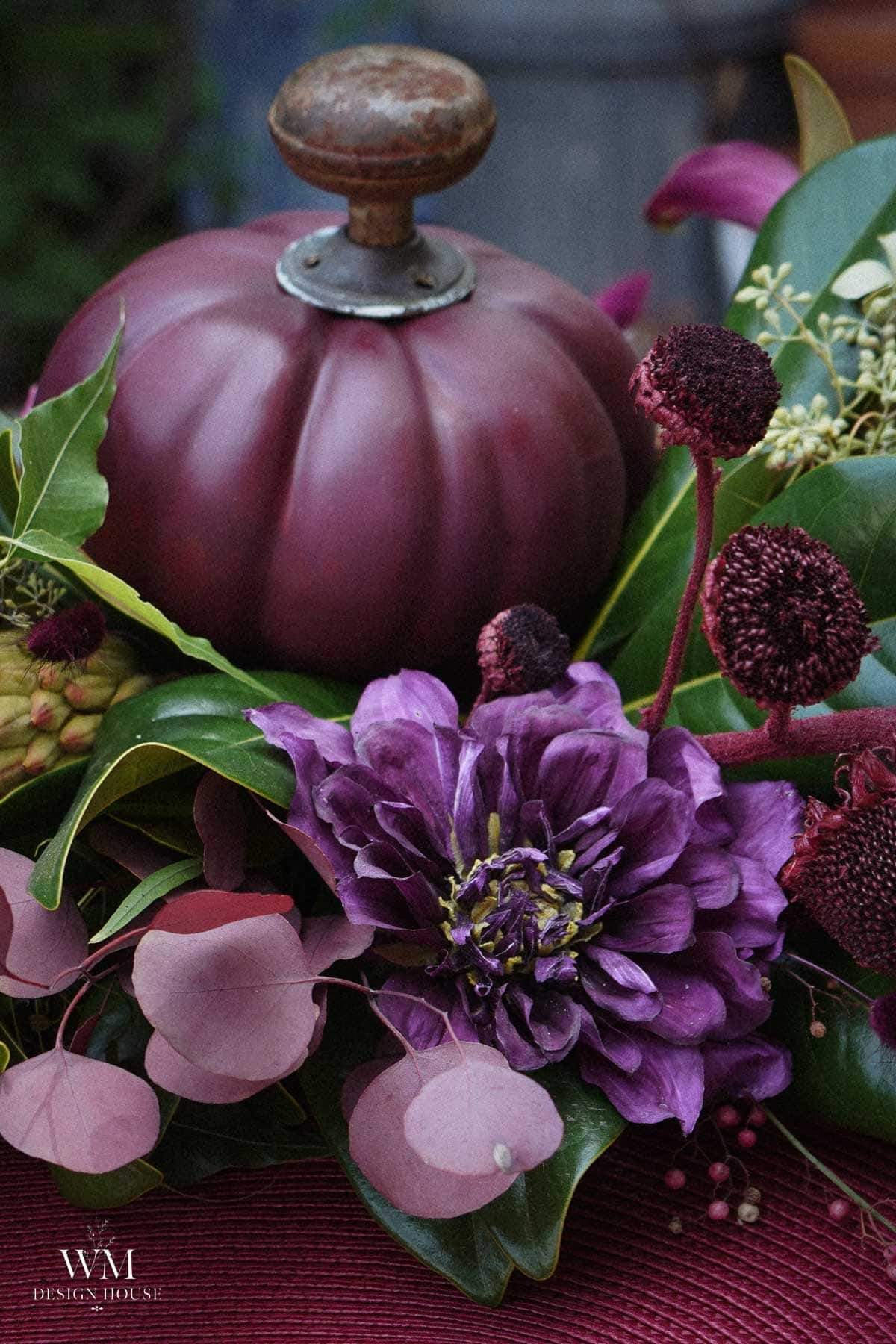 A purple pumpkin- sits among purple dried dahlias, green leaves, and reddish foliage on a farm table 
