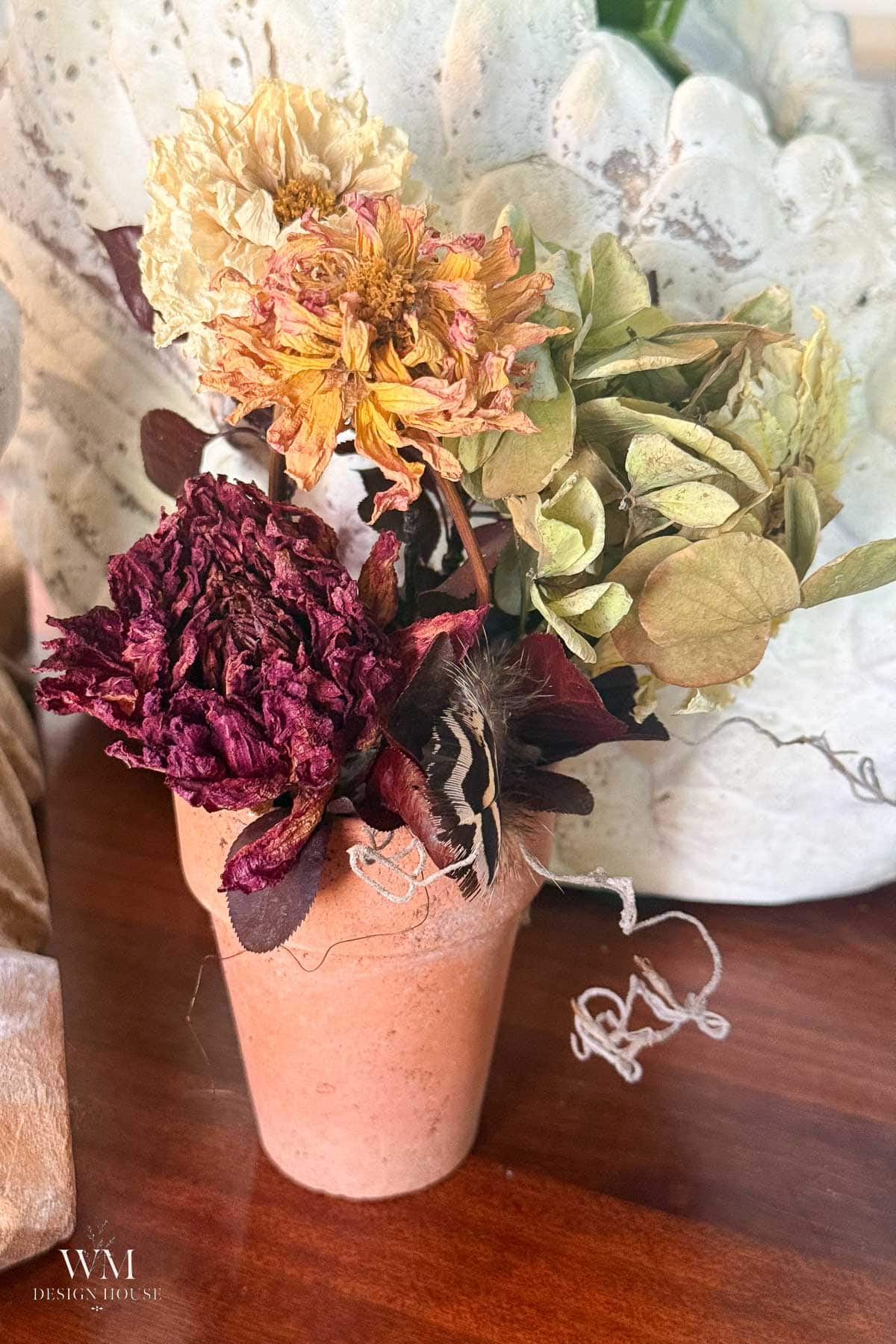 A small terracotta pot holds dried flowers in shades of purple, yellow, and green—each carefully preserved using techniques like how to dry dahlias—sitting on a wooden surface with a white decorative background.