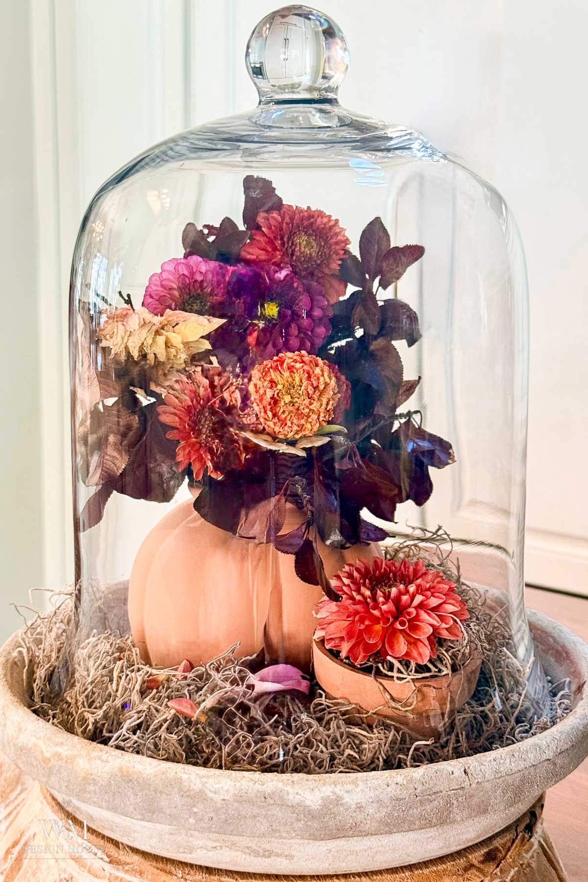 A glass cloche covers a small pumpkin vase filled with colorful flowers—perfect inspiration if you’re curious about how to dry dahlias—surrounded by moss and petals, all set on a round stone tray.